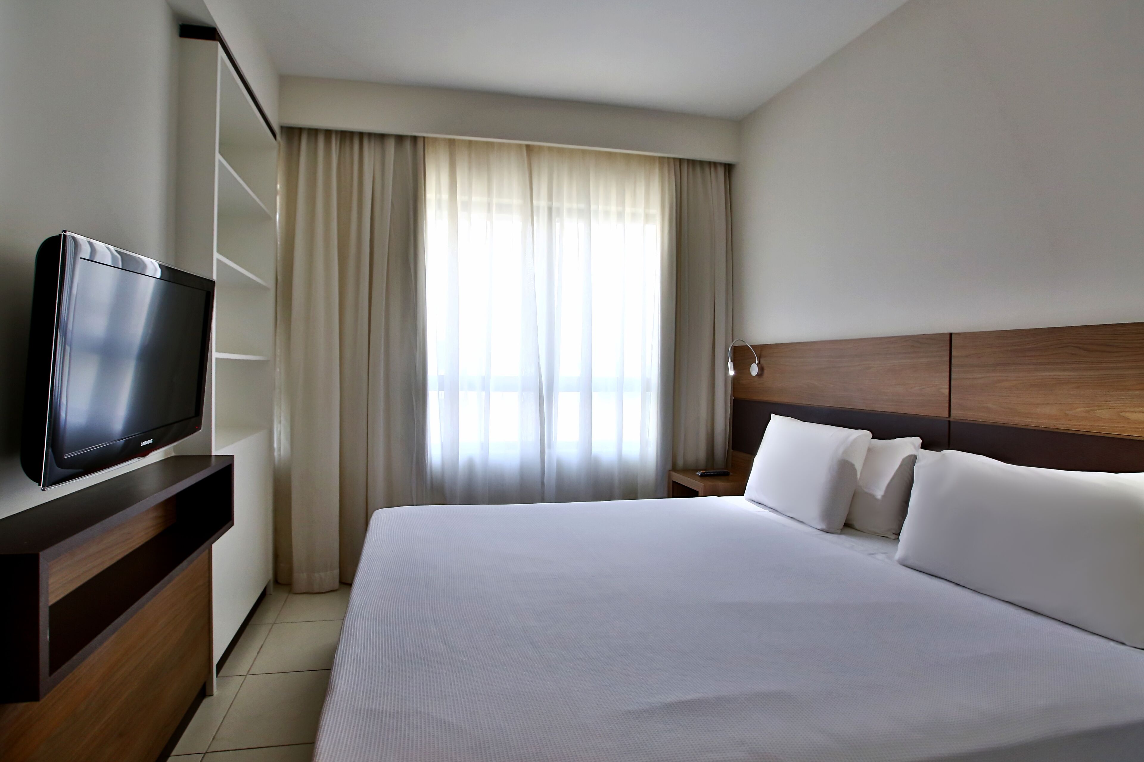Habitación de hotel con una cama grande, un televisor de pantalla plana montado sobre una consola de madera, una mesita de noche con una lámpara y una ventana con cortinas de gasa.