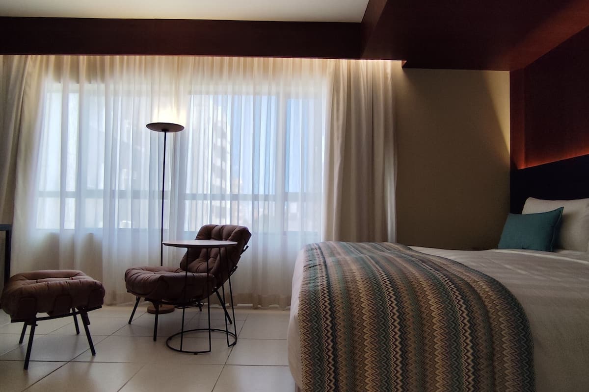 Une chambre d’hôtel avec un lit, une chaise, une petite table et une lampe sur pied.