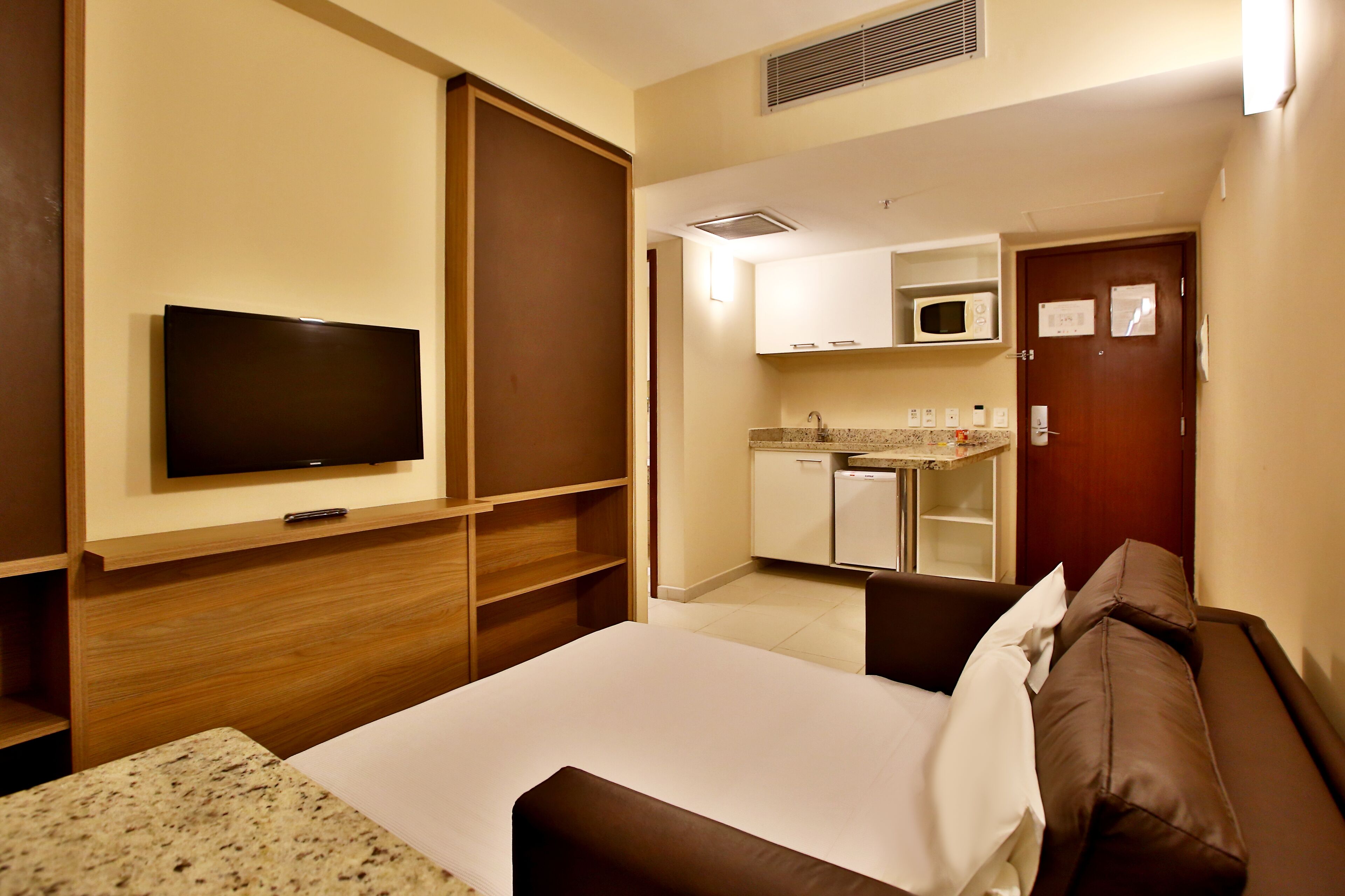 Una habitación de hotel con cama, televisor, kitchenette y una puerta.