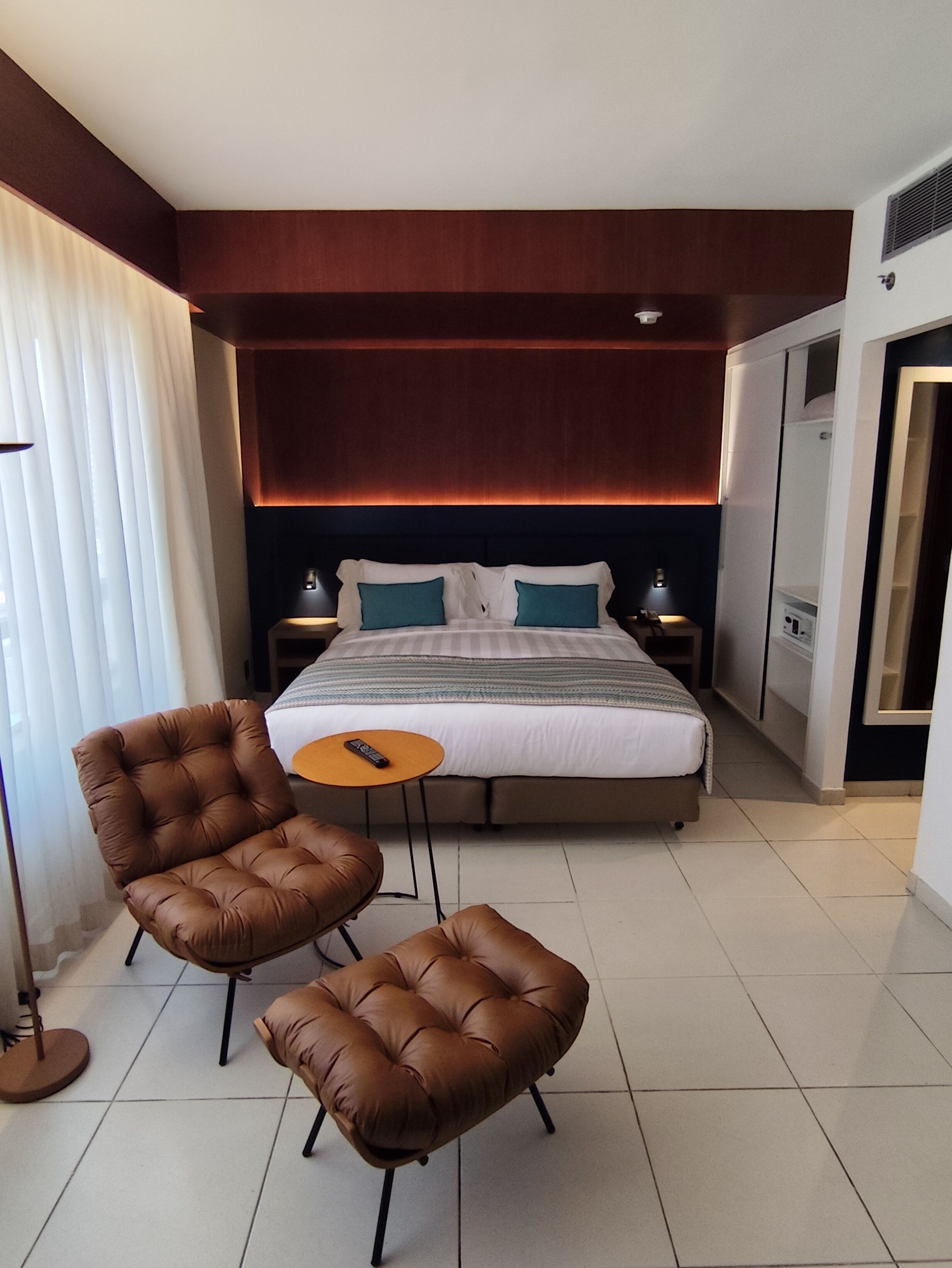Une chambre d’hôtel avec un grand lit, deux fauteuils en cuir marron, une petite table ronde et une table de chevet avec une lampe.