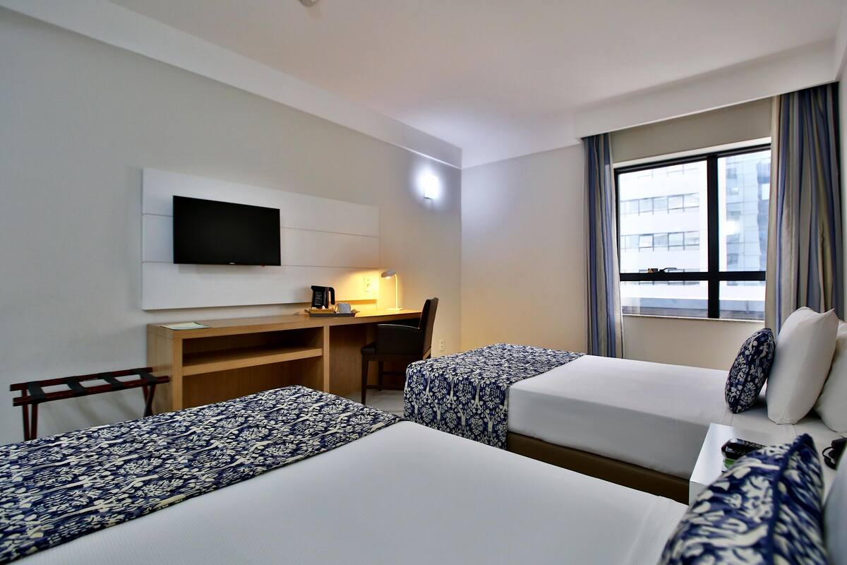 Ein Hotelzimmer mit zwei Betten, einem Schreibtisch mit Stuhl, einem Fernseher und einem Fenster mit Vorhängen.
