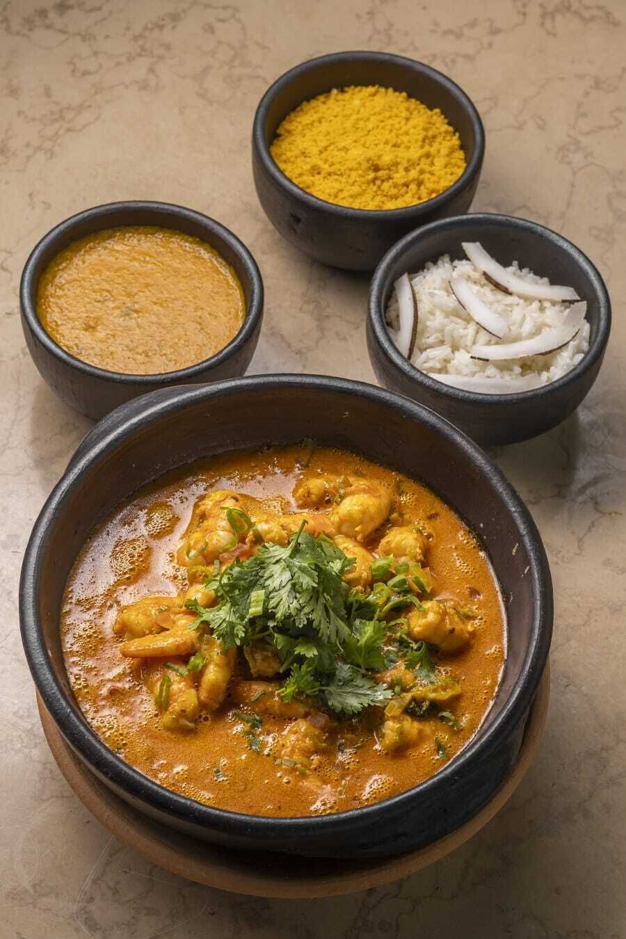 Un bol de curry au poulet, garni de coriandre, accompagné de trois accompagnements : un mélange d’épices jaunes, une poudre jaune et du riz blanc à la noix de coco.