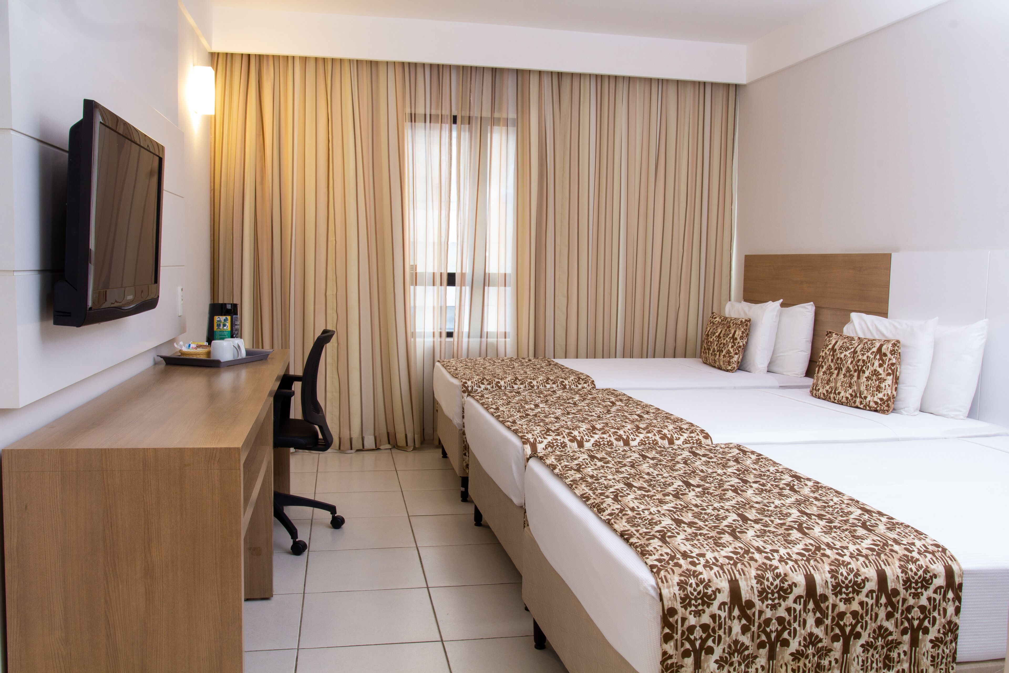 Quarto de hotel com cama, escrivaninha, cadeira, televisão e janela com cortinas.