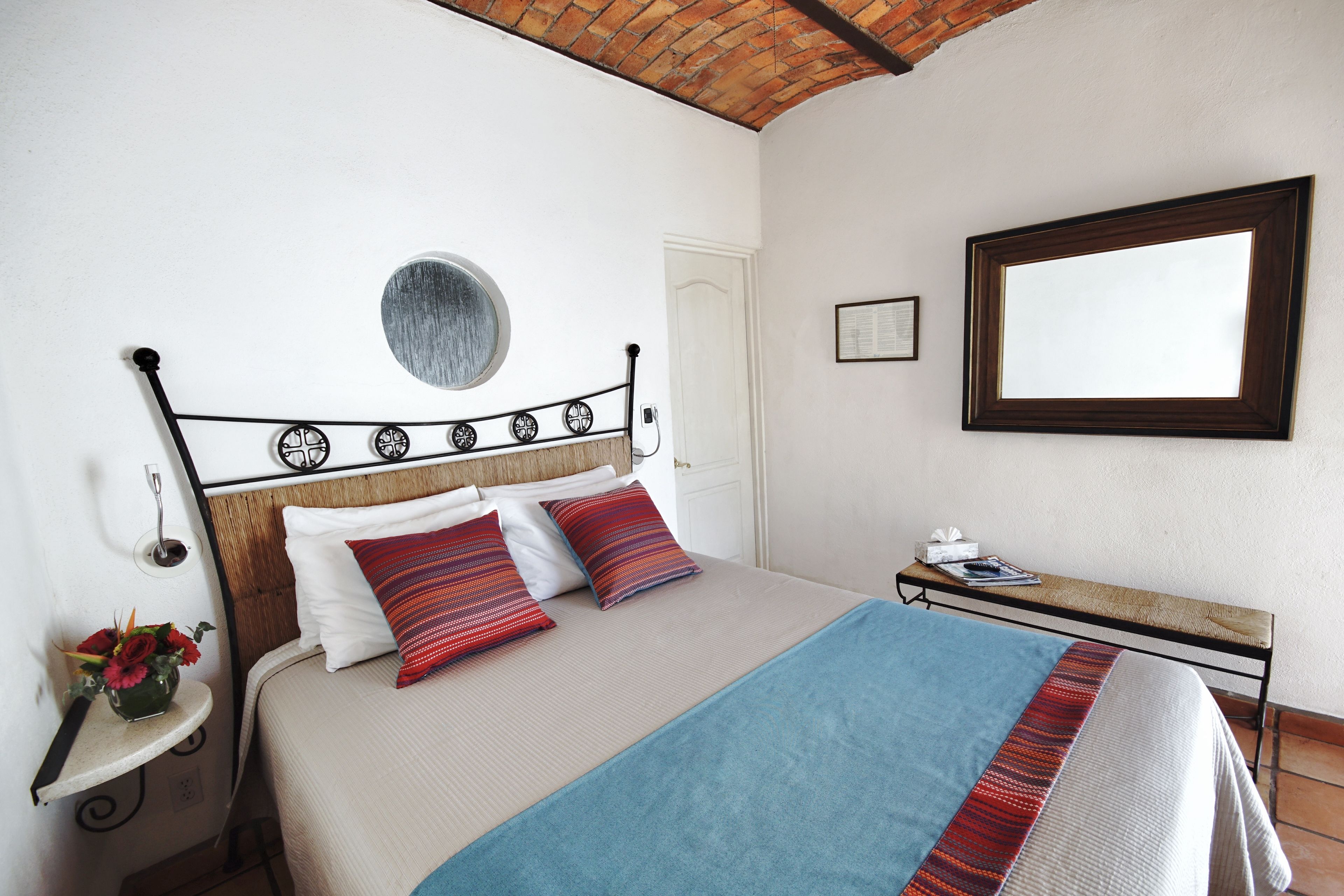 Ein ordentlich bezogenes Bett mit blauer Decke und roten Kissen, ein kleiner Nachttisch mit Blumenvase und ein an der Wand befestigter Spiegel.