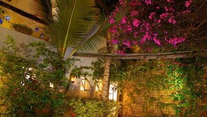 Una piscina rettangolare circondata da vegetazione lussureggiante e fiori rosa, con una palma e un edificio sullo sfondo.