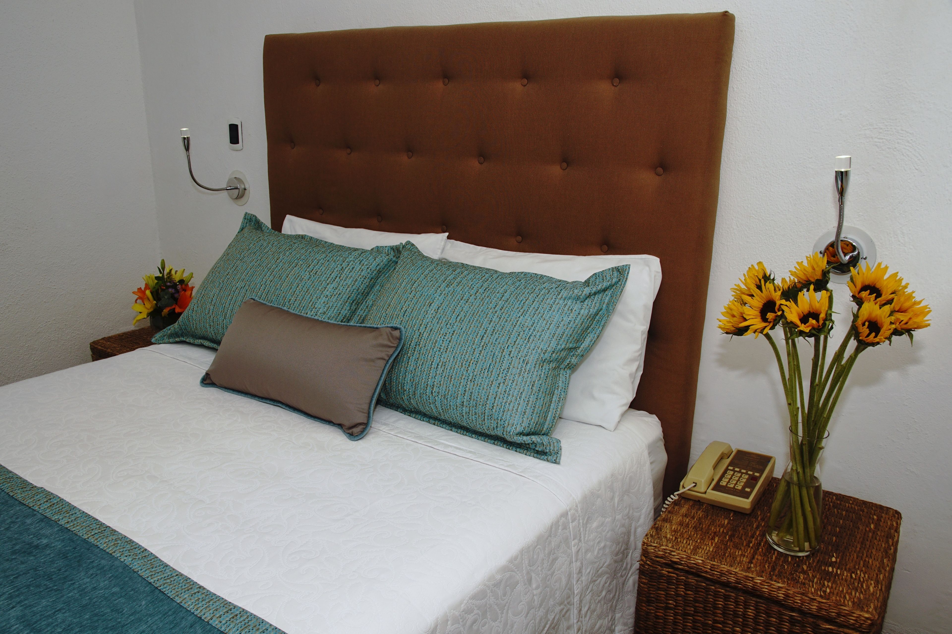 A neatly made bed with a brown headboard, a teal and brown pillow, and a white pillow. There is a wicker side table with a vase of sunflowers and a telephone.