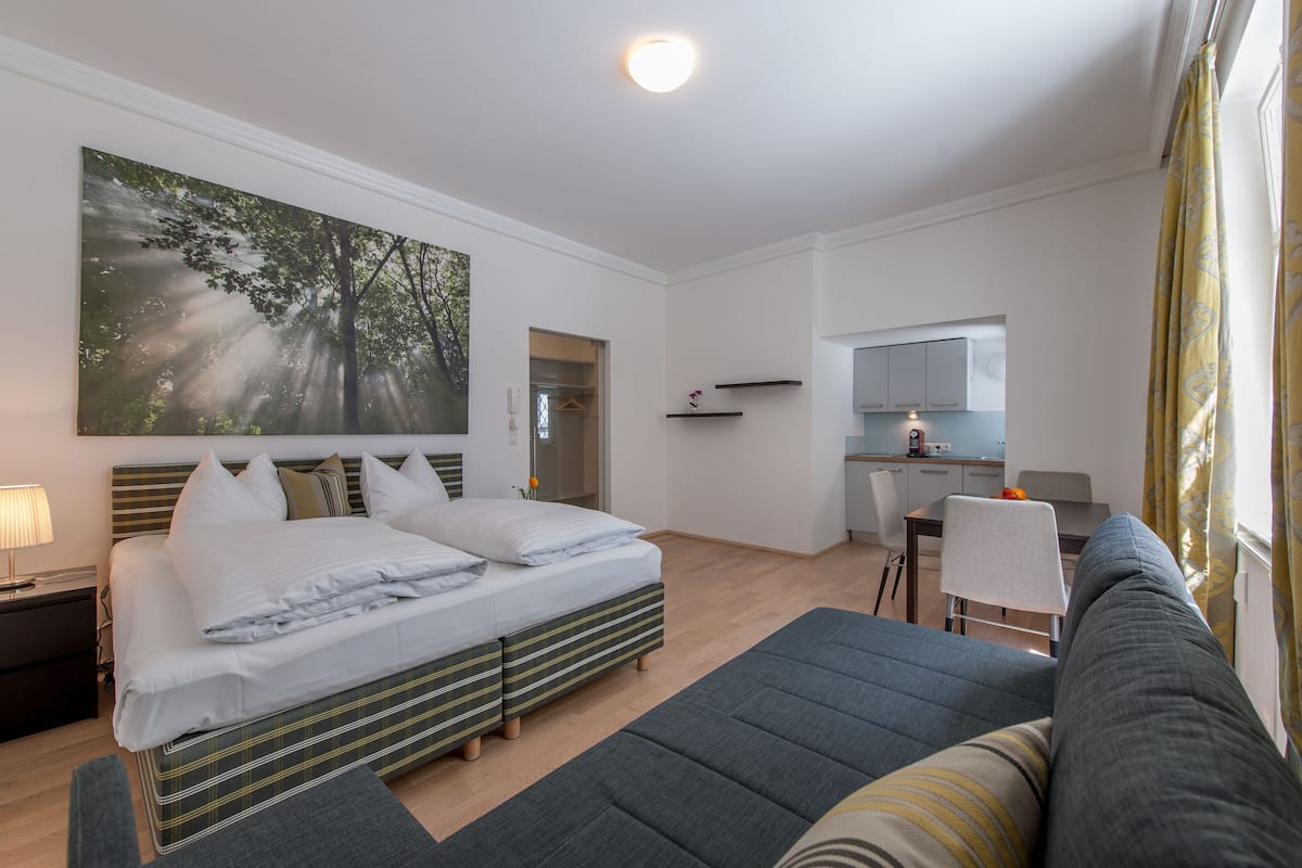 A modern hotel room with a large bed, a sofa, a dining area, and a kitchen.