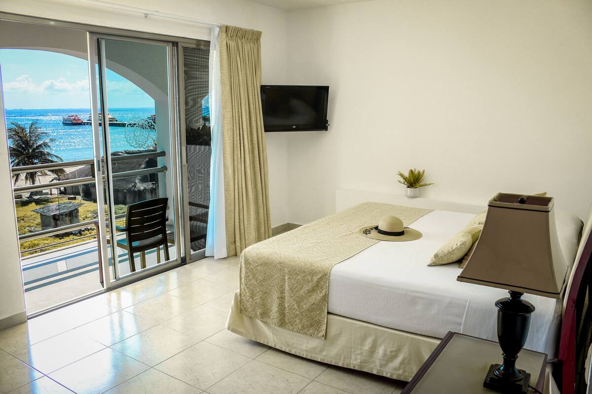 A hotel room with a bed, a TV, a lamp, a balcony with a view of the sea and boats, and a chair.