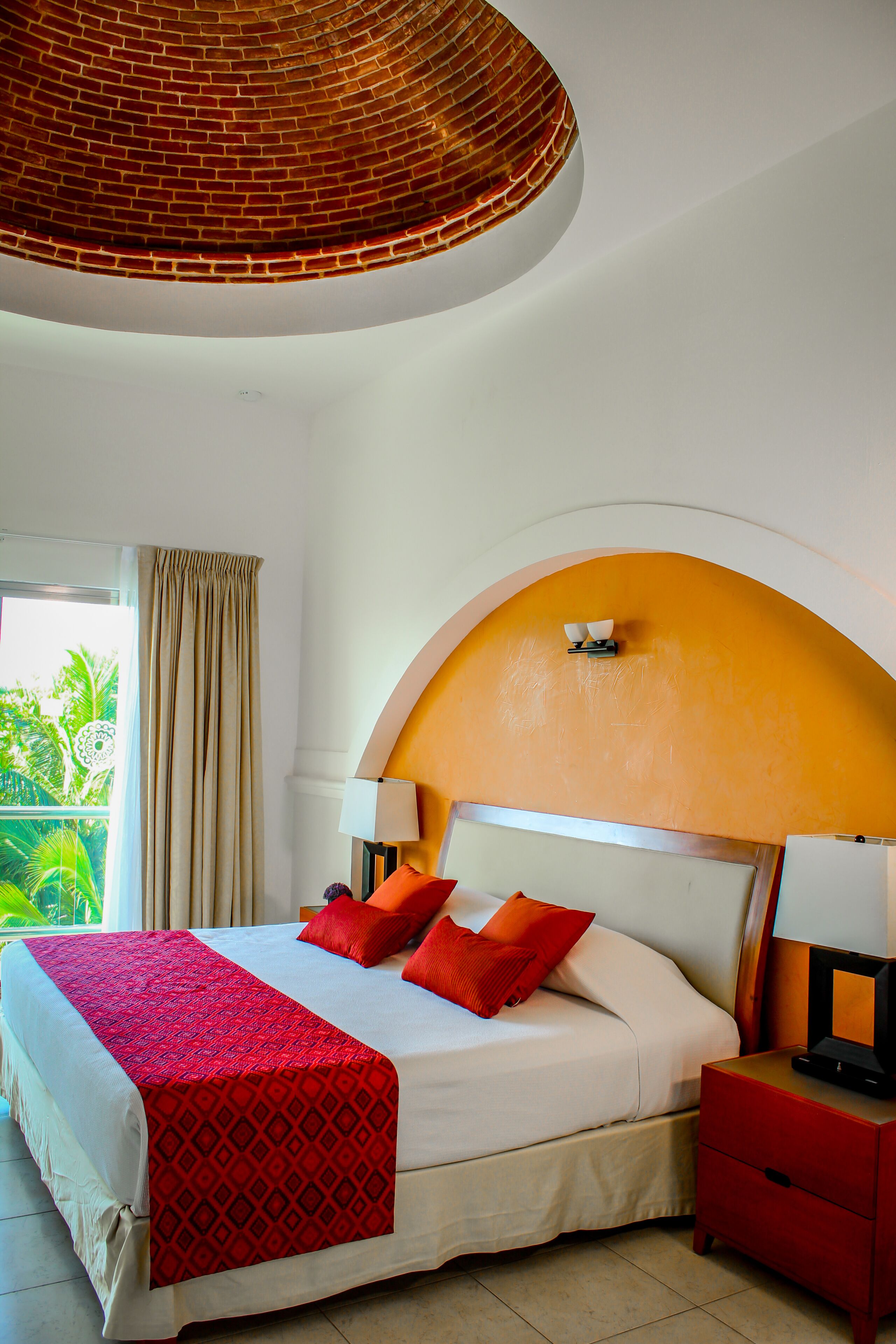 Une chambre d’hôtel avec un lit, des oreillers rouges, une table de chevet rouge, une lampe et une vue sur la verdure à l’extérieur.