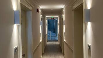 Un pasillo de hotel con una escalera central, lámparas de pared y un detector de incendios a la izquierda.