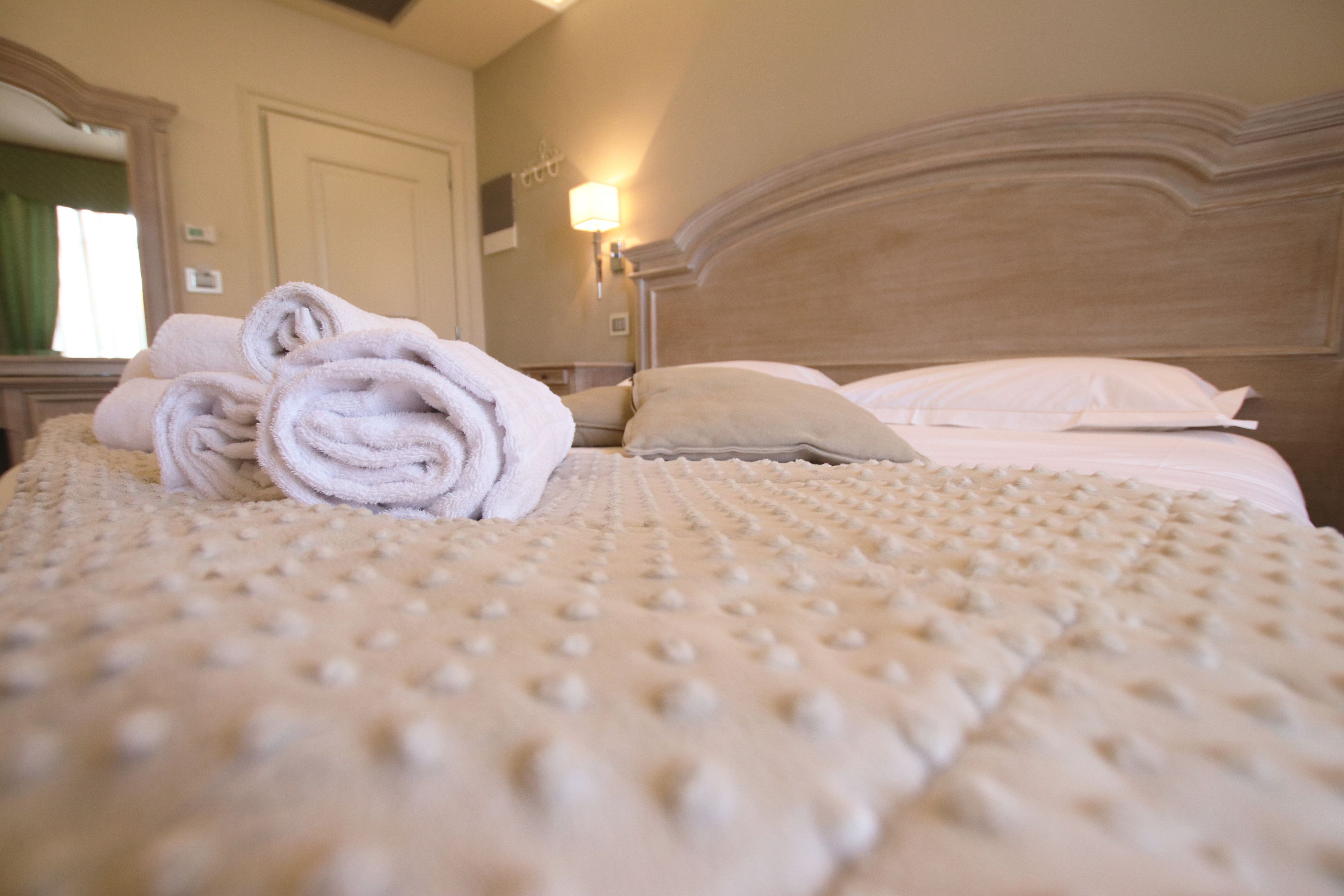 A neatly made bed with rolled towels, a textured bedspread, and a headboard with a light fixture.