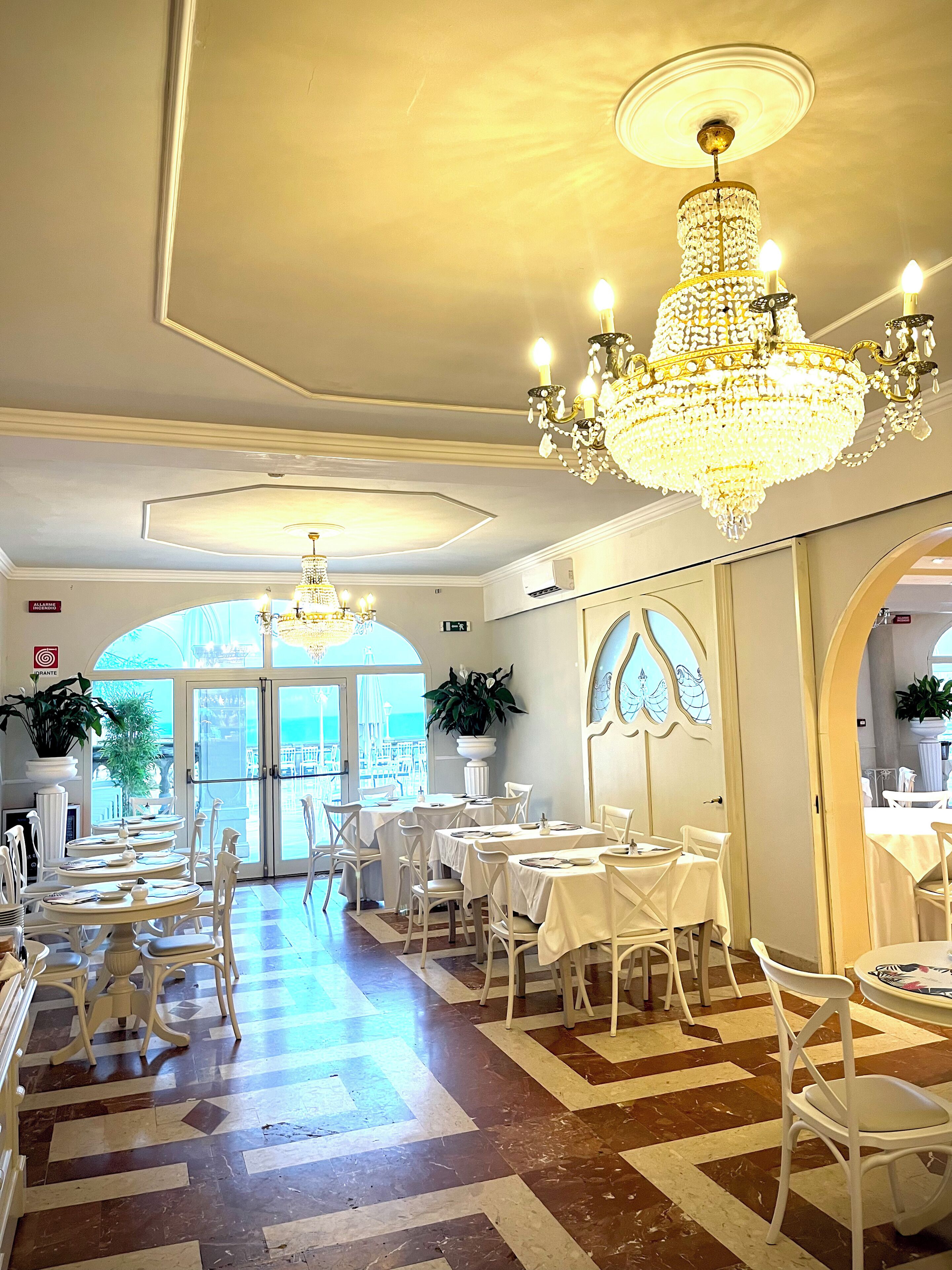 A dining area with white tables and chairs, a large chandelier, and a view of the sea through the glass doors.
