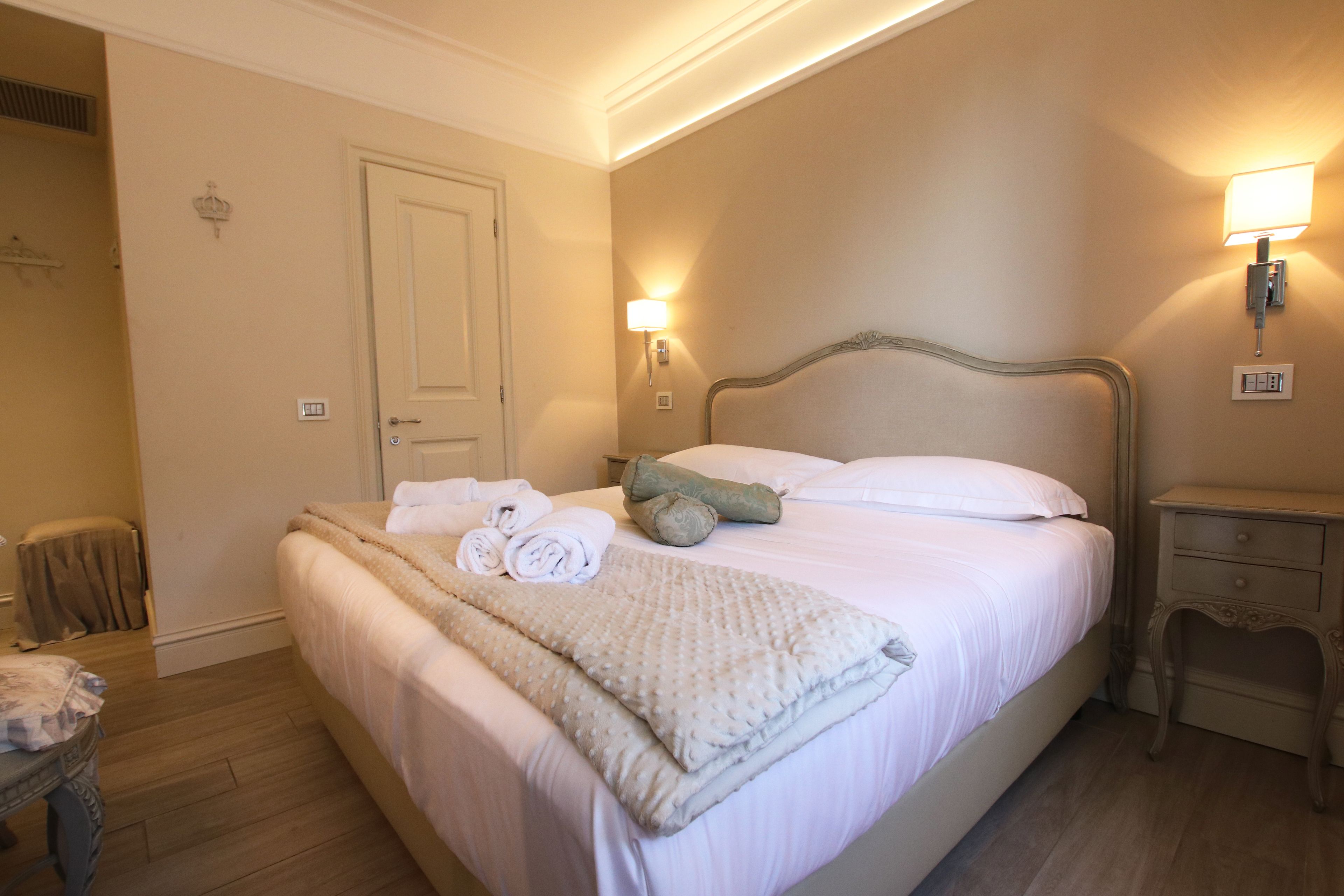 Un lit bien fait, avec du linge de lit blanc et une couverture beige, une table de chevet avec une lampe, et une porte visible au fond.