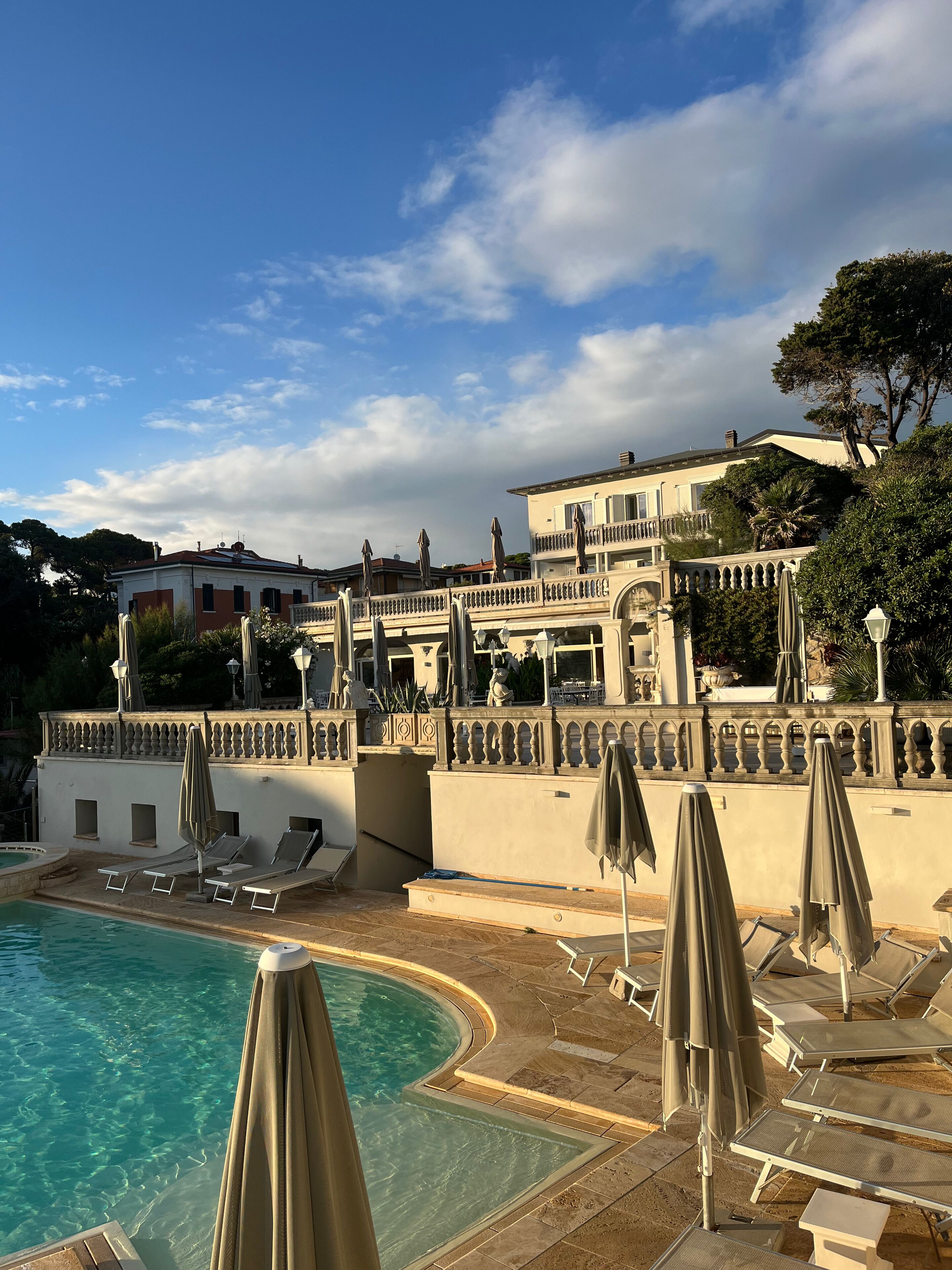 Un espace piscine avec des chaises longues et des parasols, un bâtiment doté de balcons et d’une fontaine, et un ciel dégagé.