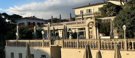 Zona de piscina con sillones y sombrillas, un edificio con balcones y una fuente, bajo un cielo despejado.