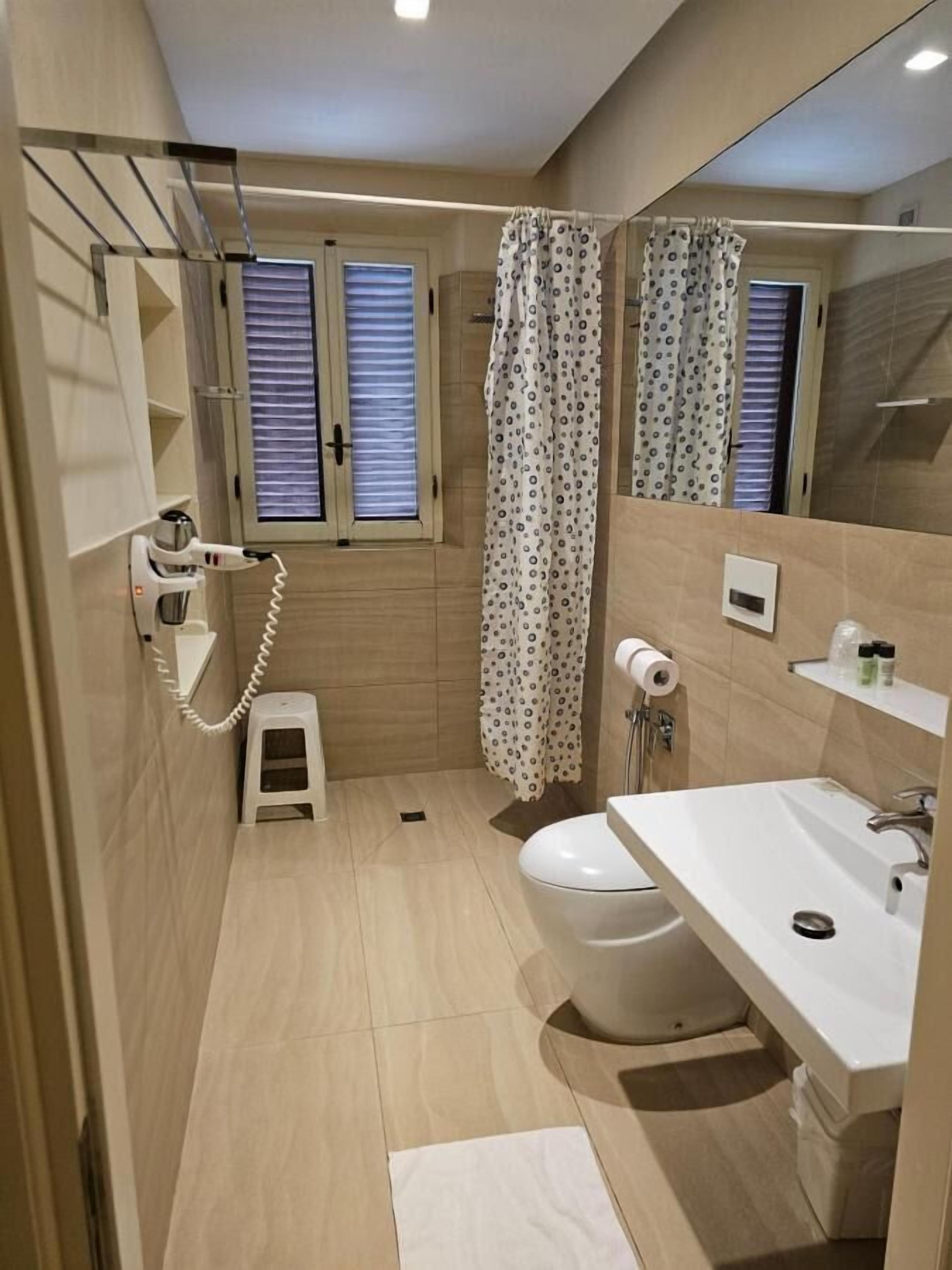 Un bagno piccolo ma funzionale, con il water, il lavandino e la doccia. Si trova uno sgabello, un asciugacapelli e un porta asciugamani.