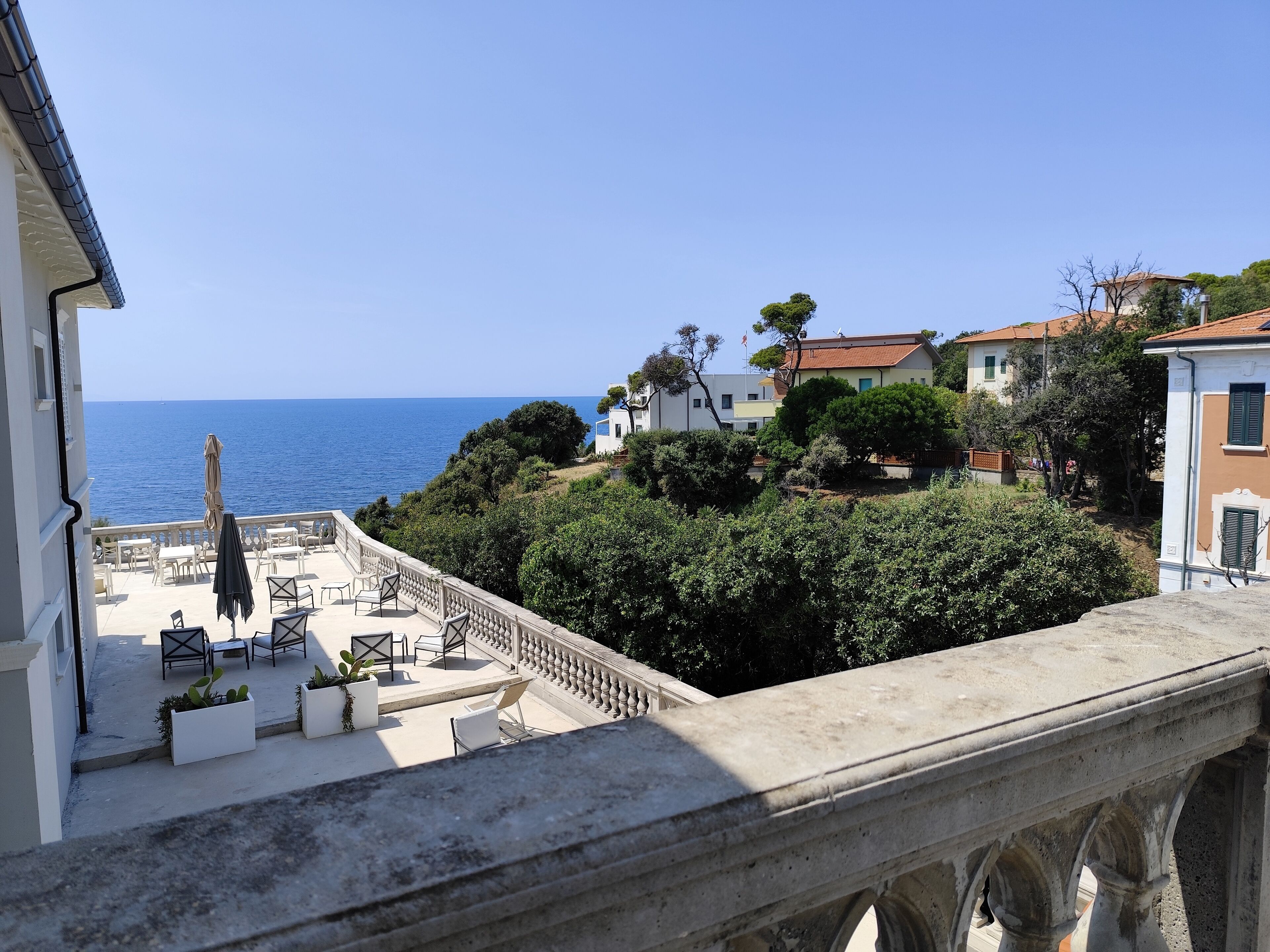 A terrace with outdoor seating, overlooking a coastal area with buildings and trees.