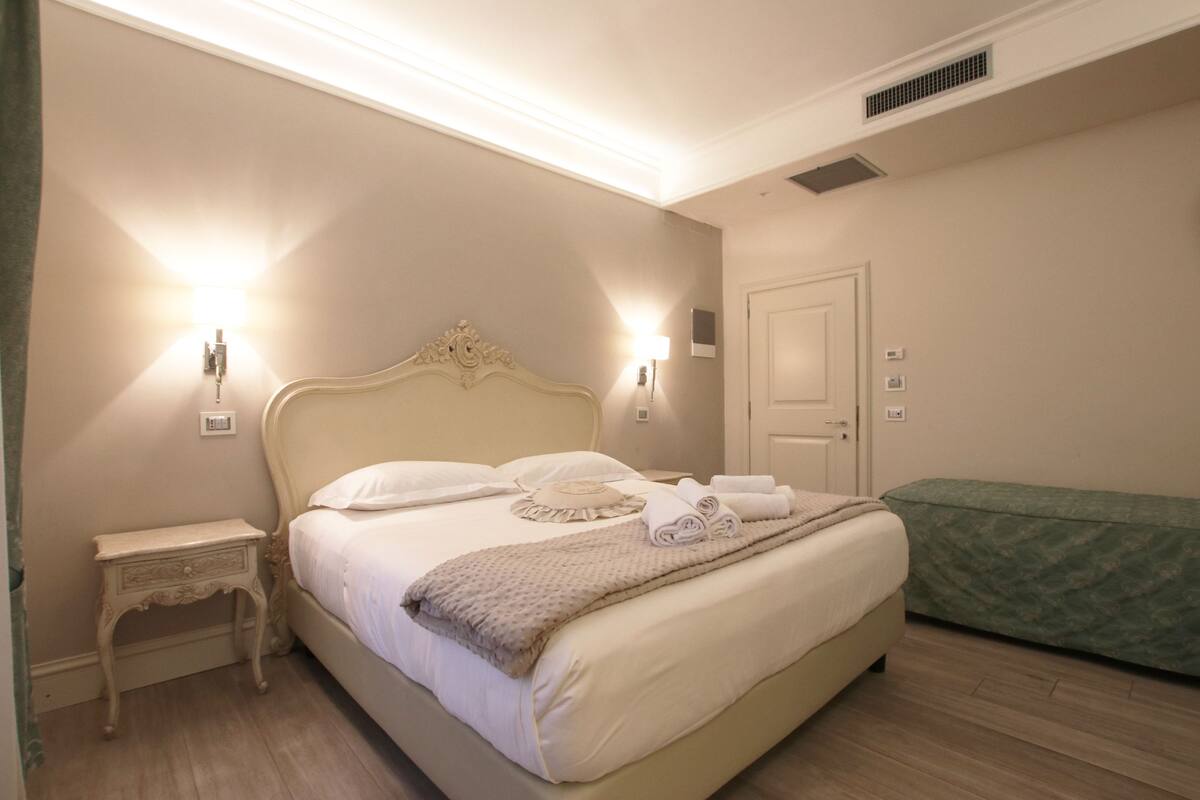 A neatly made bed with a headboard, two bedside lamps, a bedside table, and a green upholstered bench.