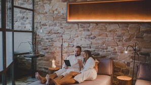 A couple relaxing on a cushioned bench in a spa-like setting with stone walls and a warm light fixture above. - Kunzmann's Hotel (Bad Bocklet)