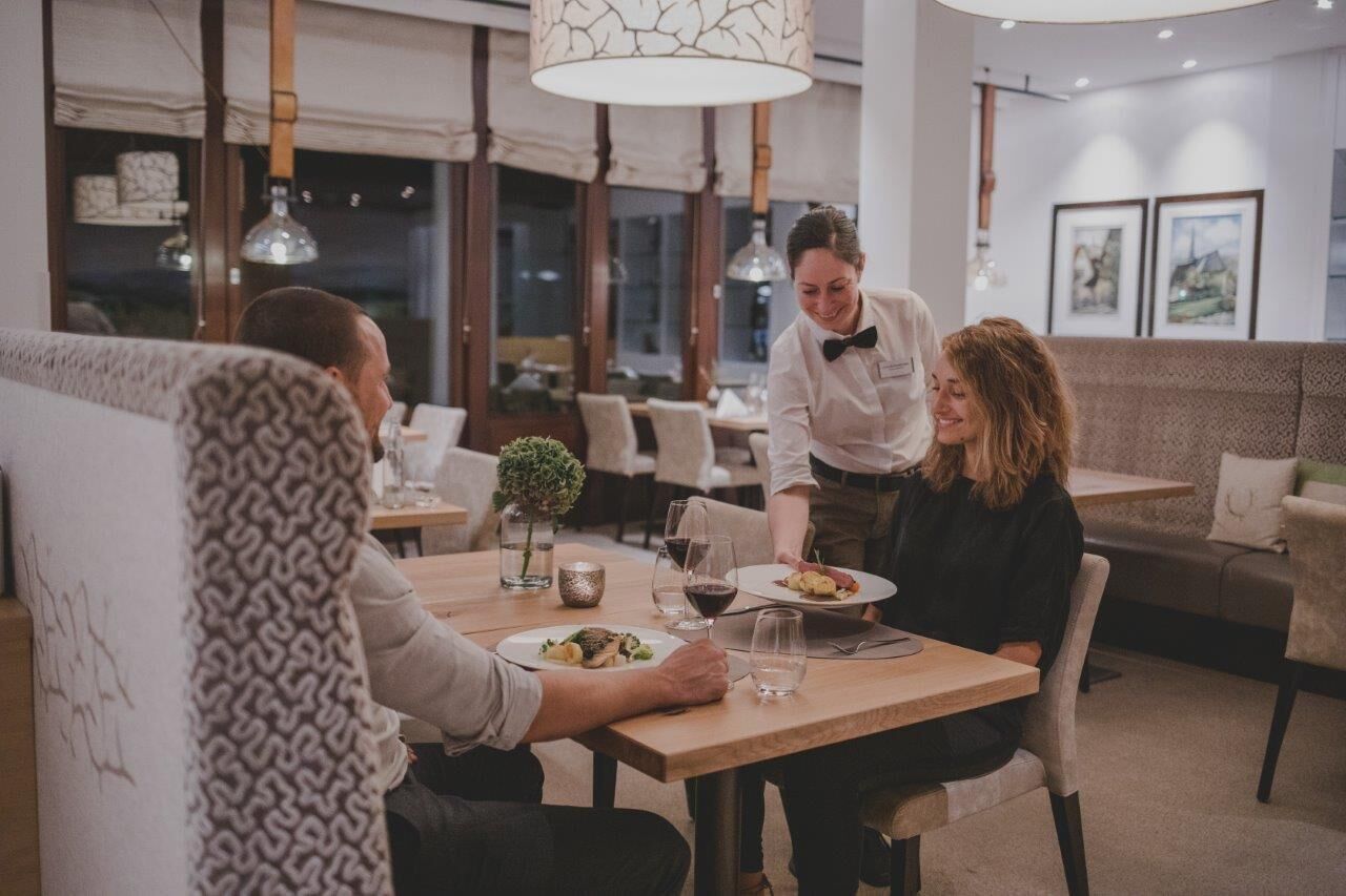 Un restaurante con mesa de madera, un camarero atendiendo a los clientes y un rincón de mesas acogedor.