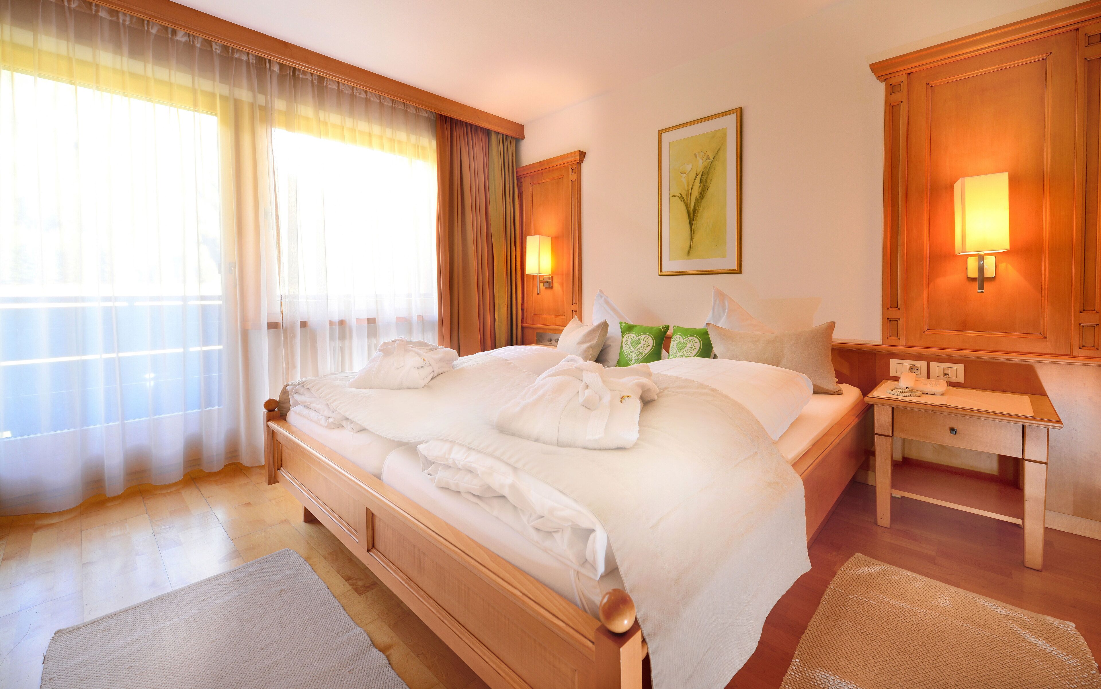Habitación de hotel con una cama grande, cabecero de madera, dos lámparas de noche, una mesita de noche y una ventana con cortinas traslúcidas.