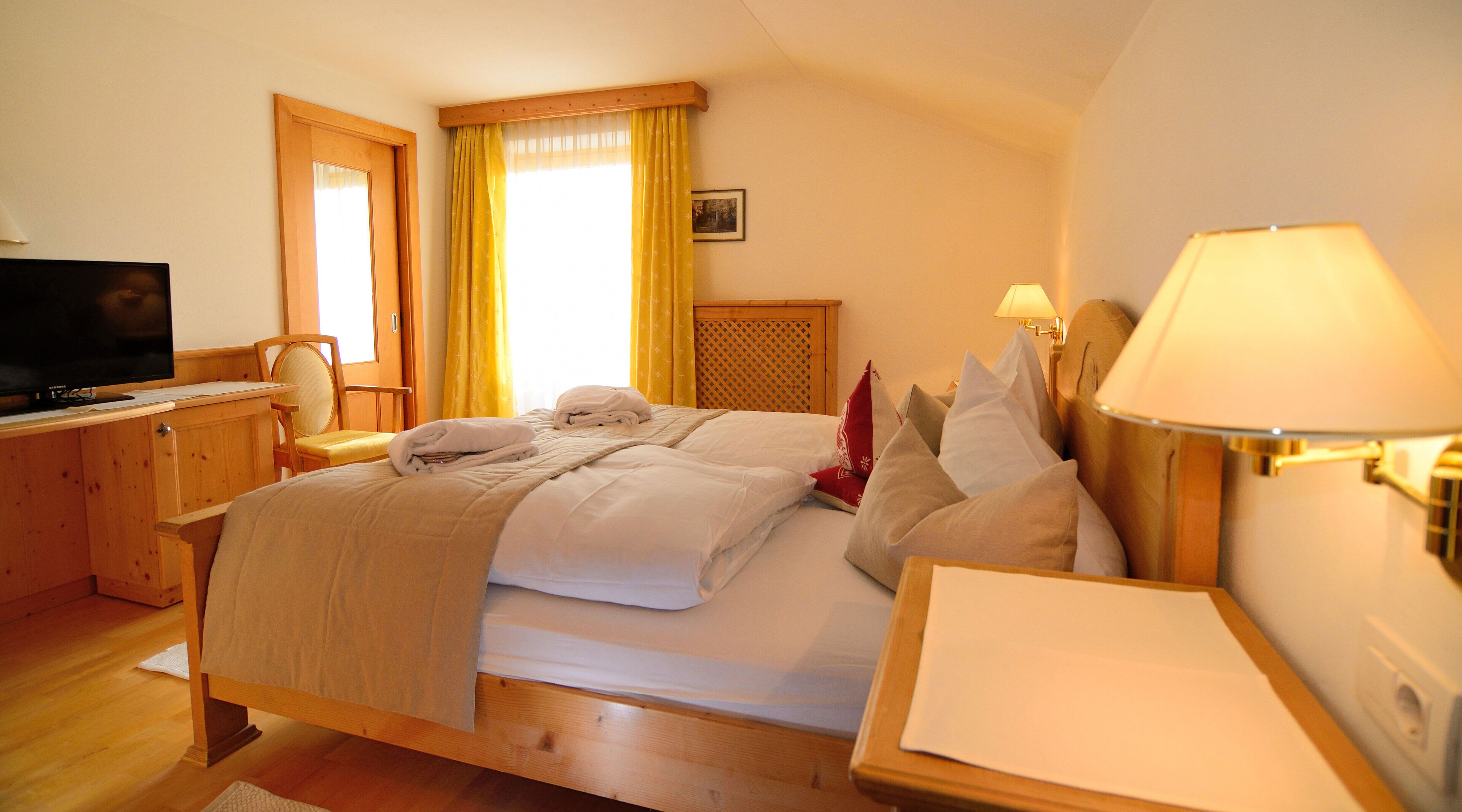 A hotel room with a wooden bed, a desk with a television, a bedside lamp, and a window with curtains.