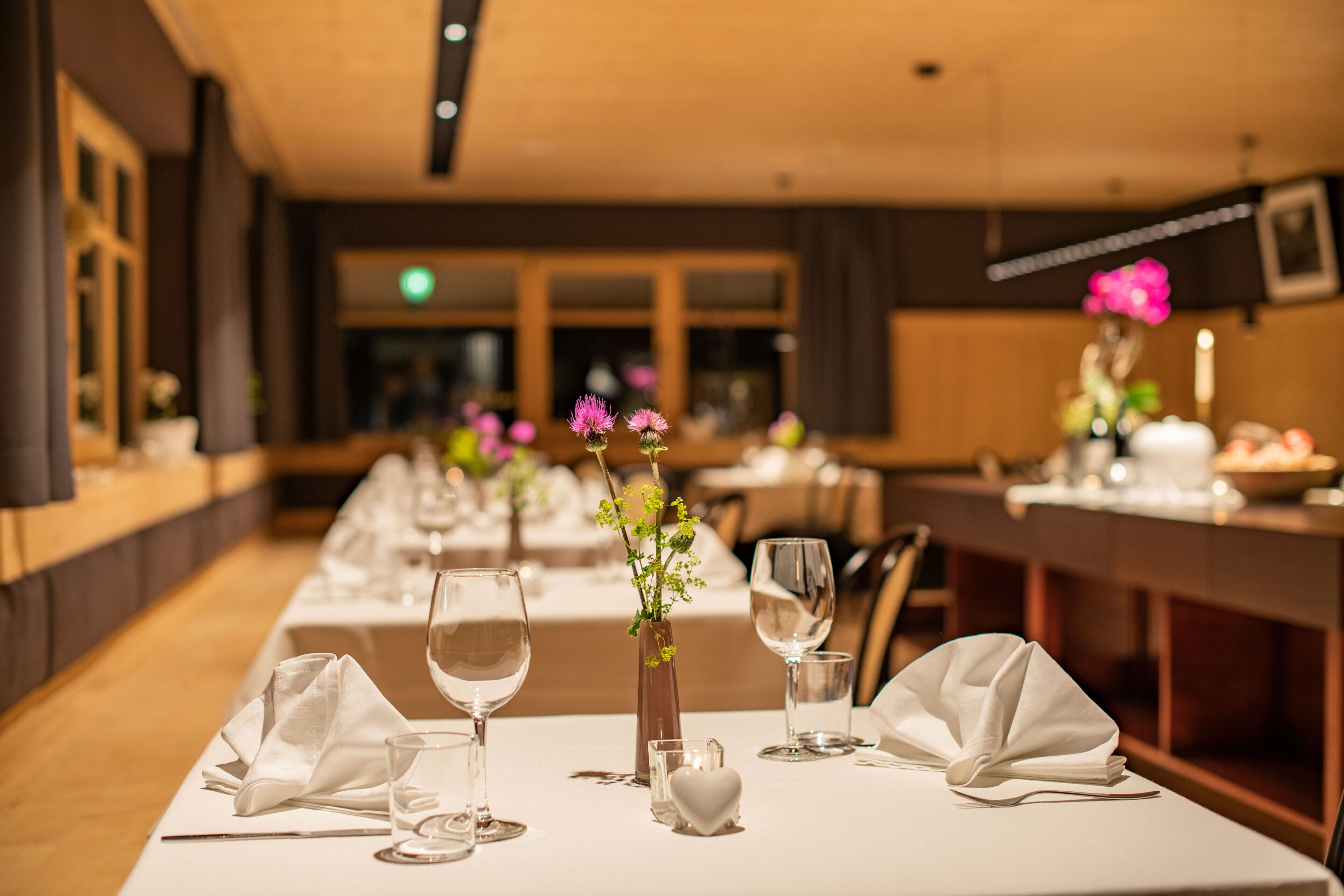 A restaurant with long tables set for dining, featuring white tablecloths, wine glasses, and small vases with flowers.