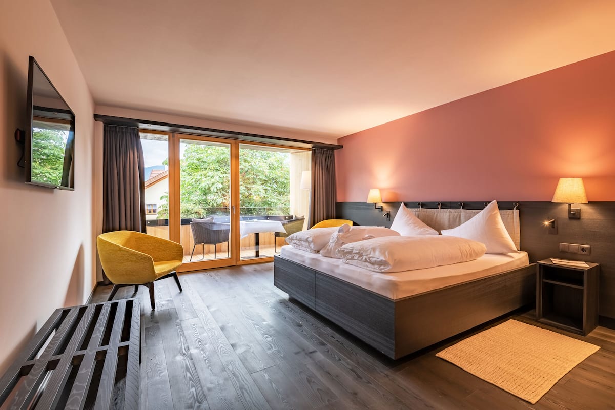 A modern hotel room with a large bed, a yellow armchair, a small table, and a view of the outdoors through a sliding glass door.