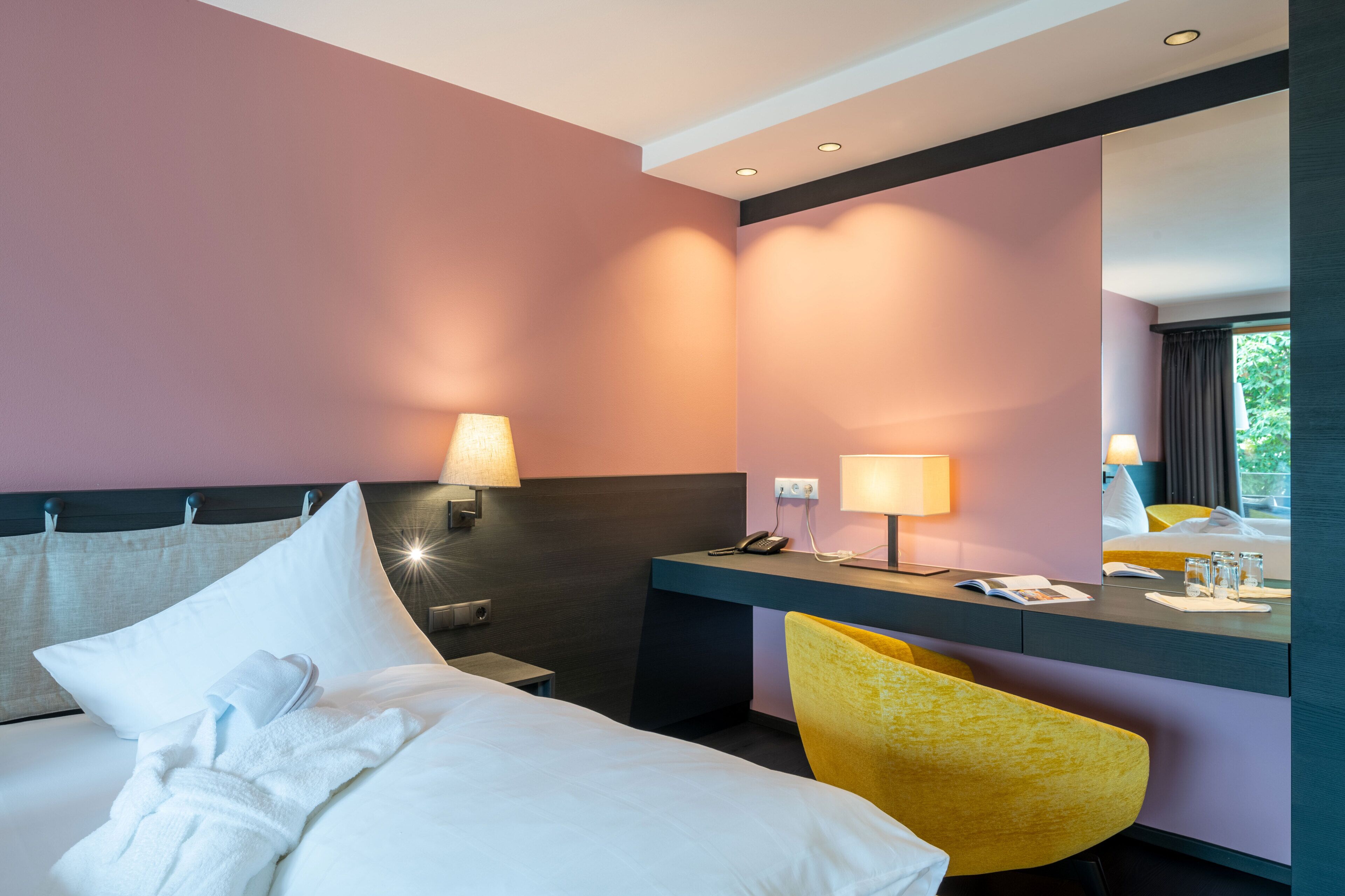 A hotel room with a bed, bedside table, lamp, chair, and a desk with a phone and glasses.