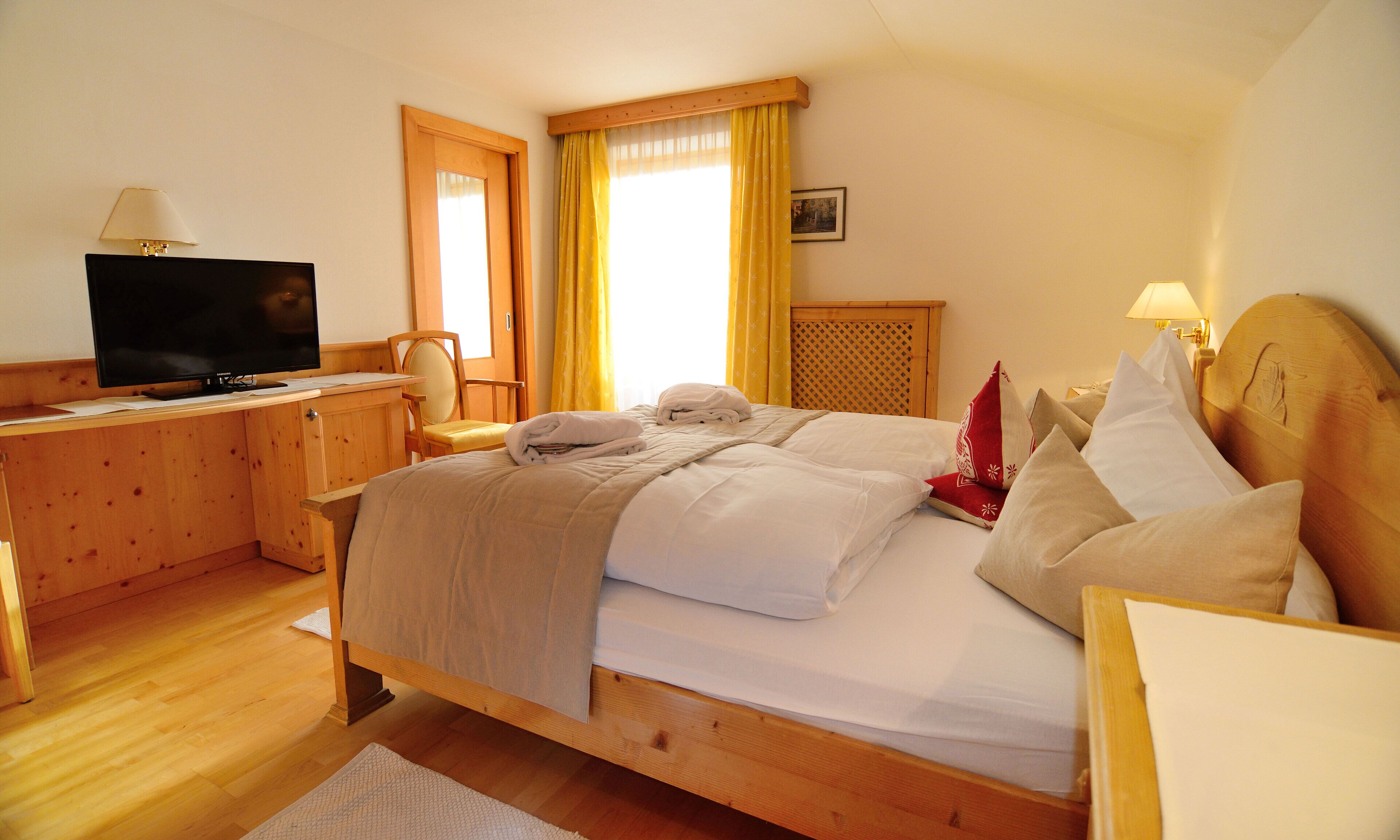 Camera d'albergo con un letto in legno, una scrivania con televisore, una sedia e una finestra con tende.