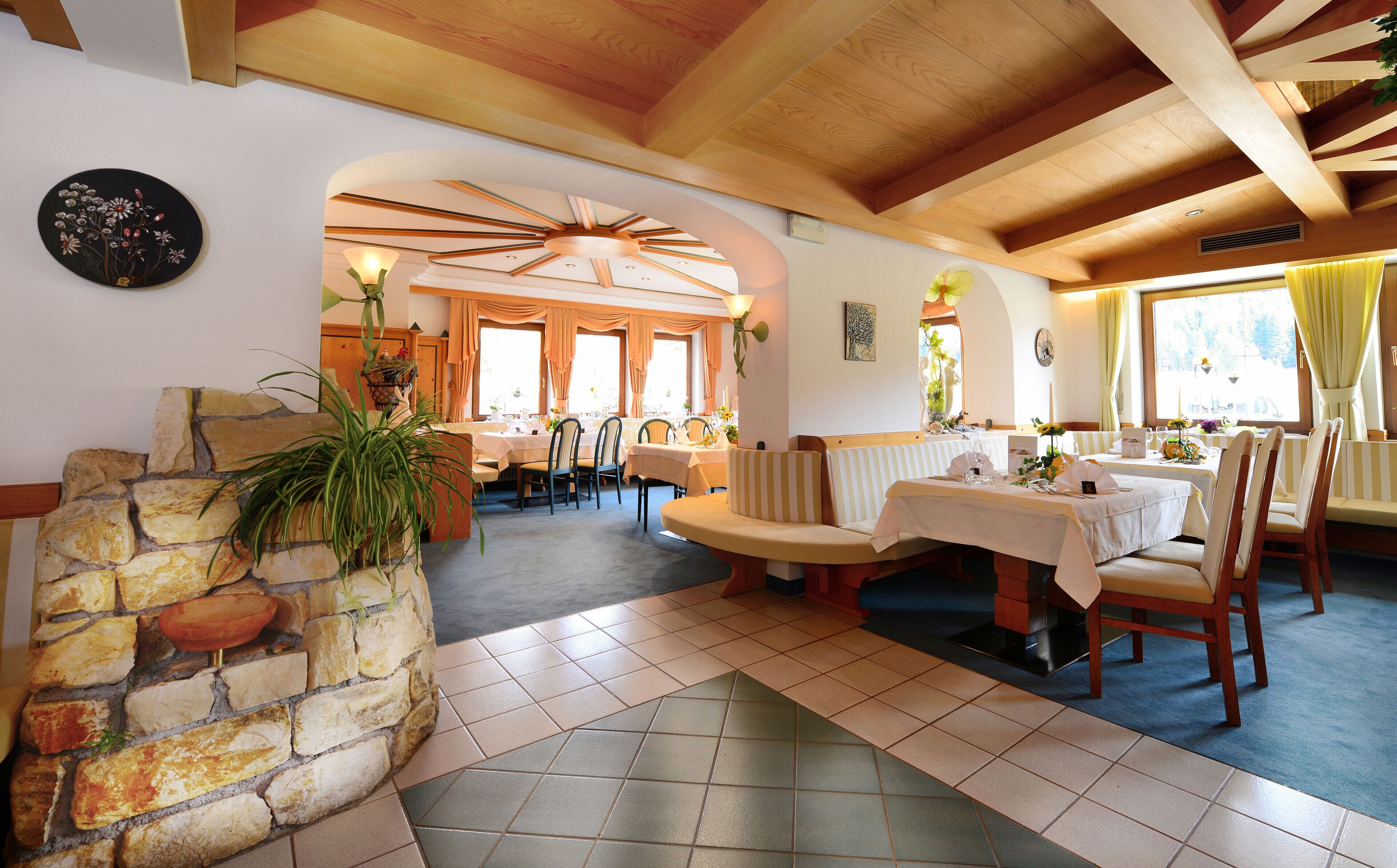 Un comedor con paredes de piedra, vigas de madera y chimenea de piedra. Las mesas están preparadas con sillas, y la habitación tiene ventanas amplias.