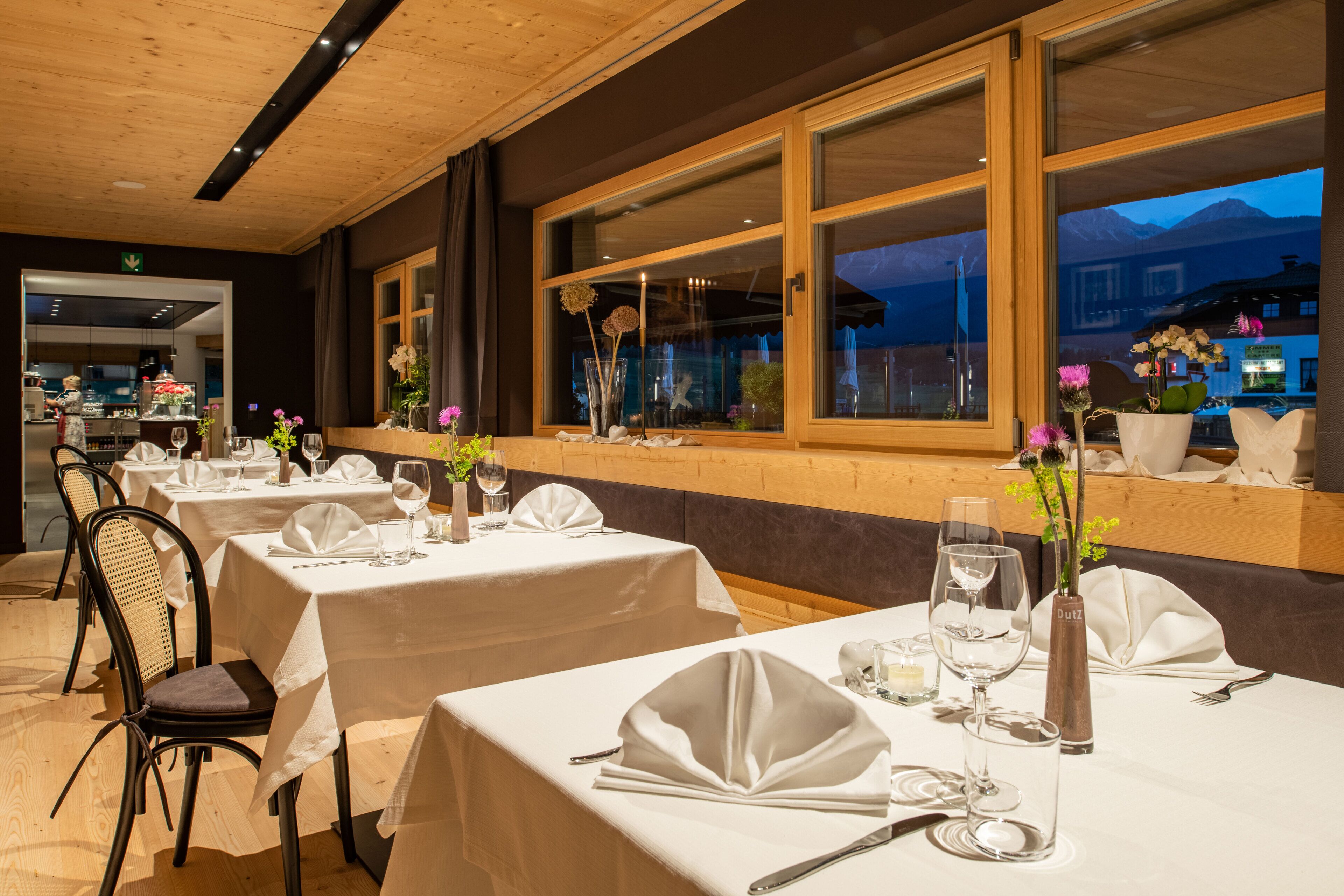A restaurant with tables set for dining, large windows with a view of mountains, and a wooden ceiling.