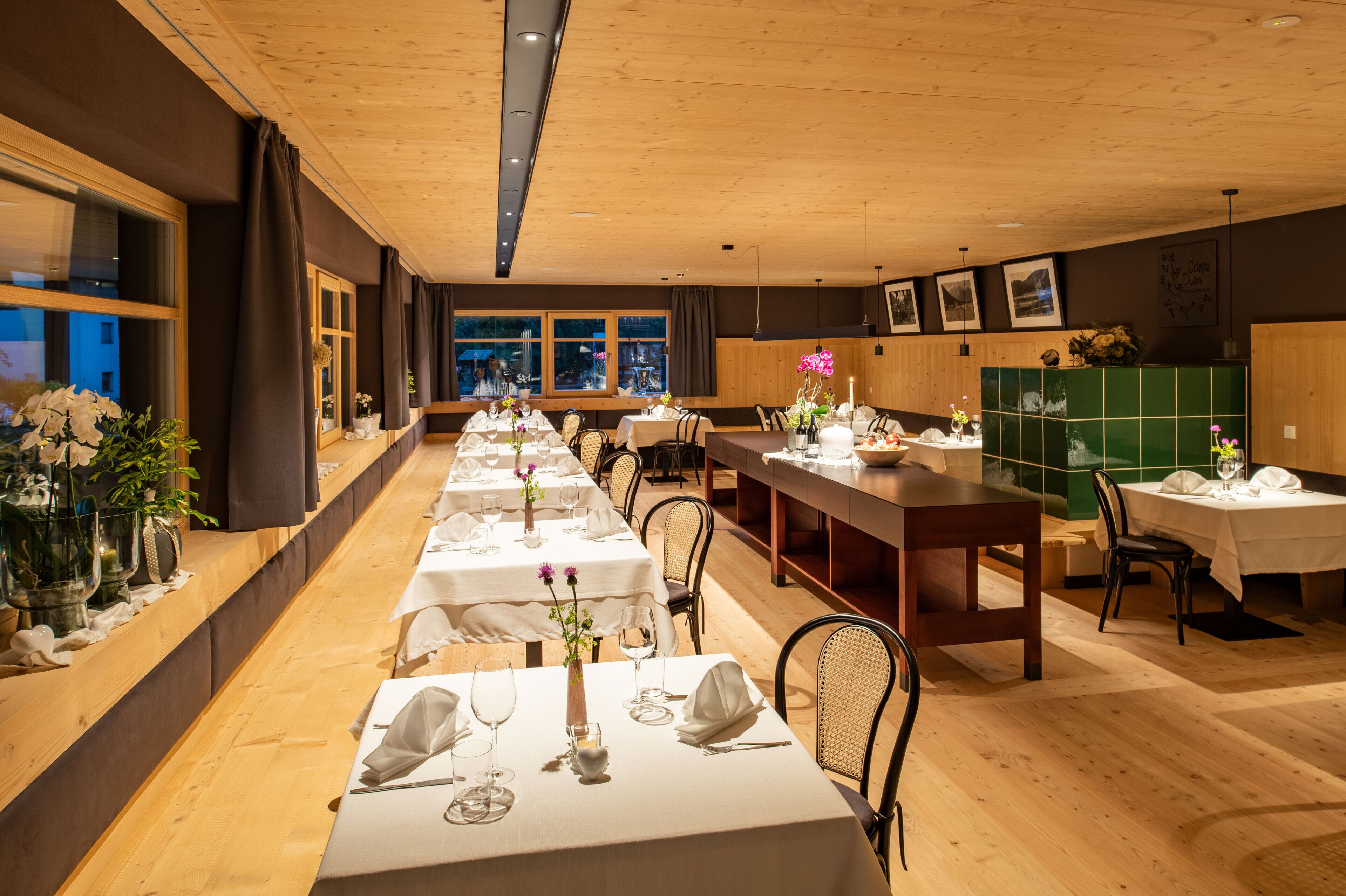 Un restaurant amb taules llargues preparades per dinar, terres de fusta i grans finestres amb cortines.