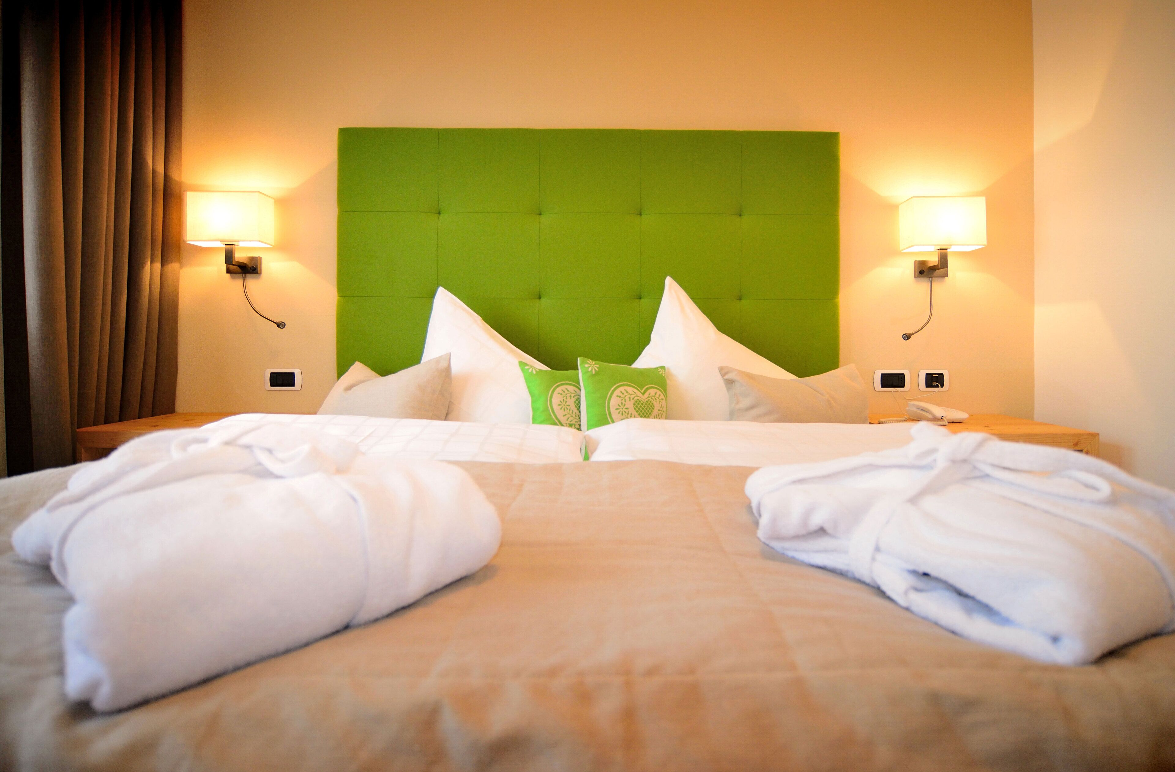 Un lit bien fait, avec du linge de lit blanc, une tête de lit recouverte d’un tissu vert et deux lampes de chevet.