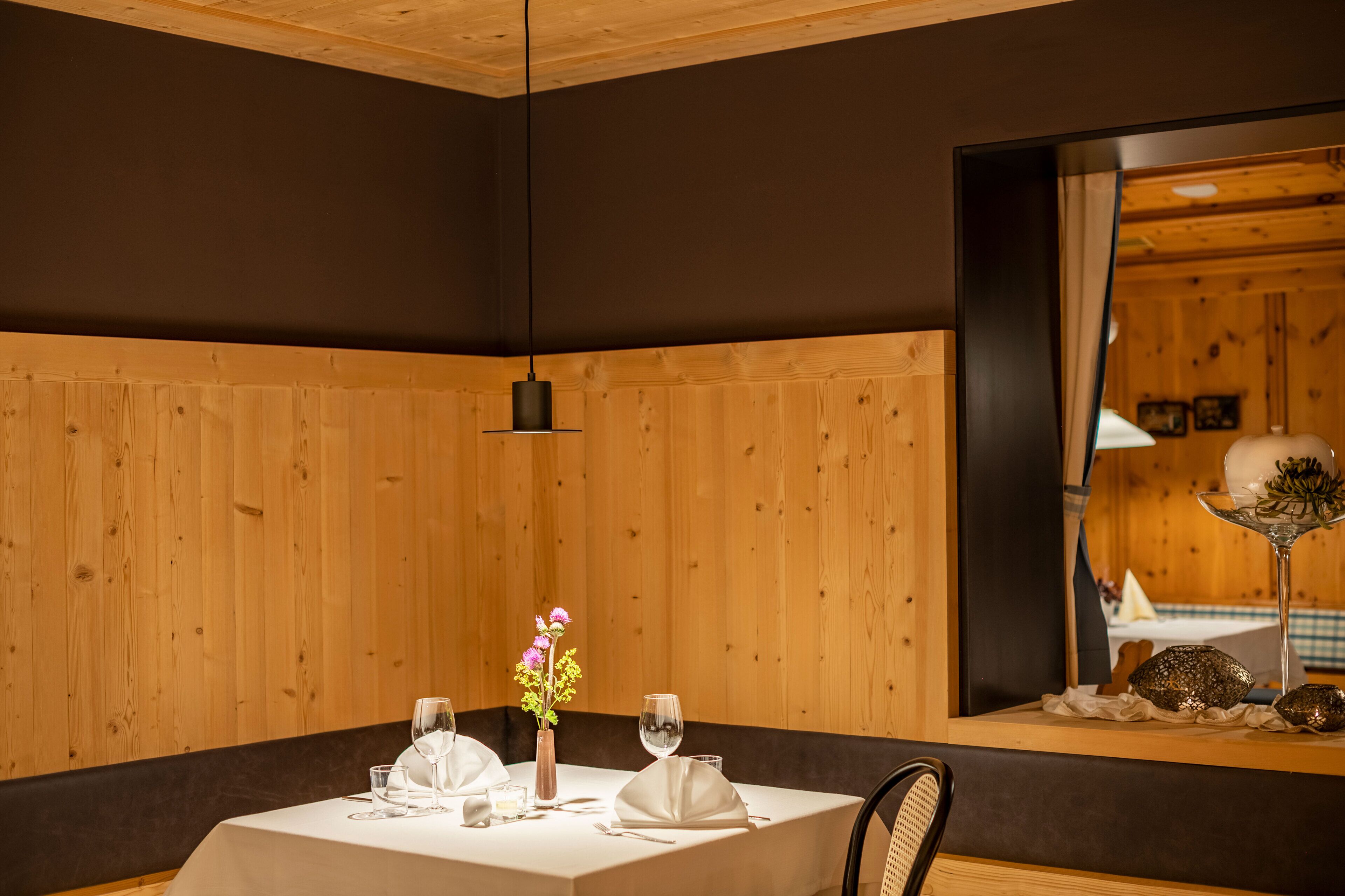 Een eetruimte met een wand bekleed met hout, een tafel gedekt met witte tafelkleden, glazen en een vaas met bloemen.