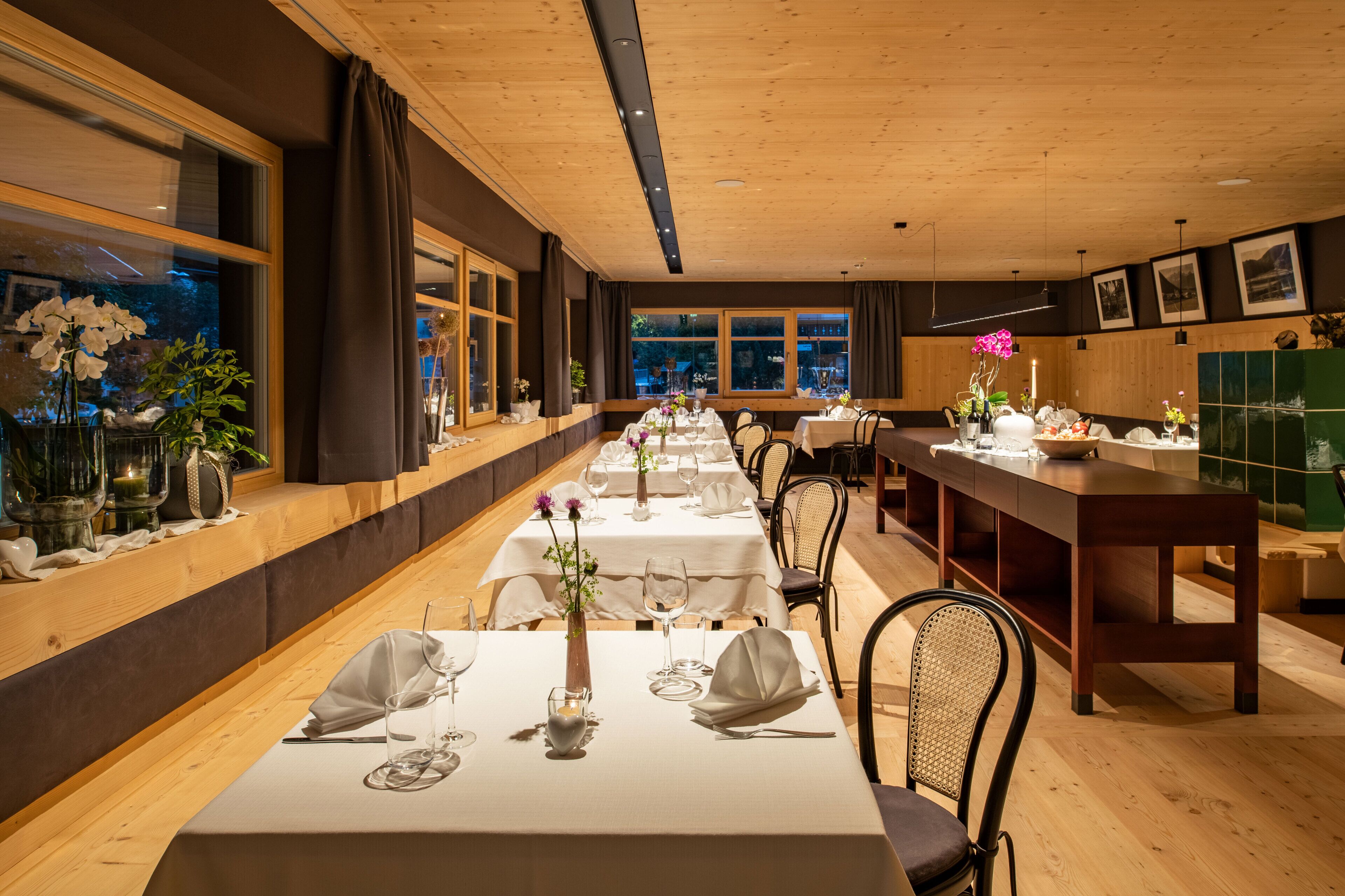 A restaurant with long tables set for dining, wooden paneling, and large windows with curtains.