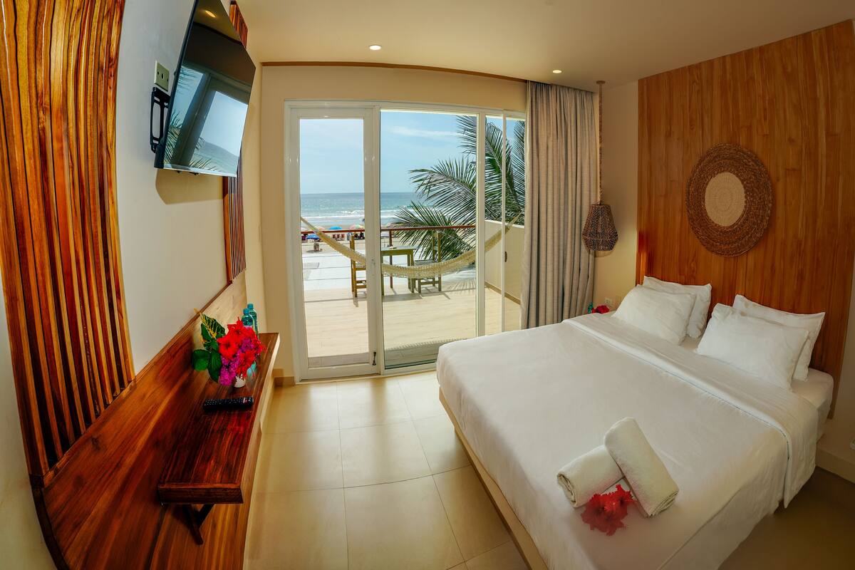 Habitación de hotel con cama, televisión, pared de madera, vista a la playa y balcón.