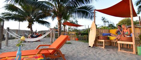 A beach setting with lounge chairs, a hammock, and wooden seating under a shaded area.