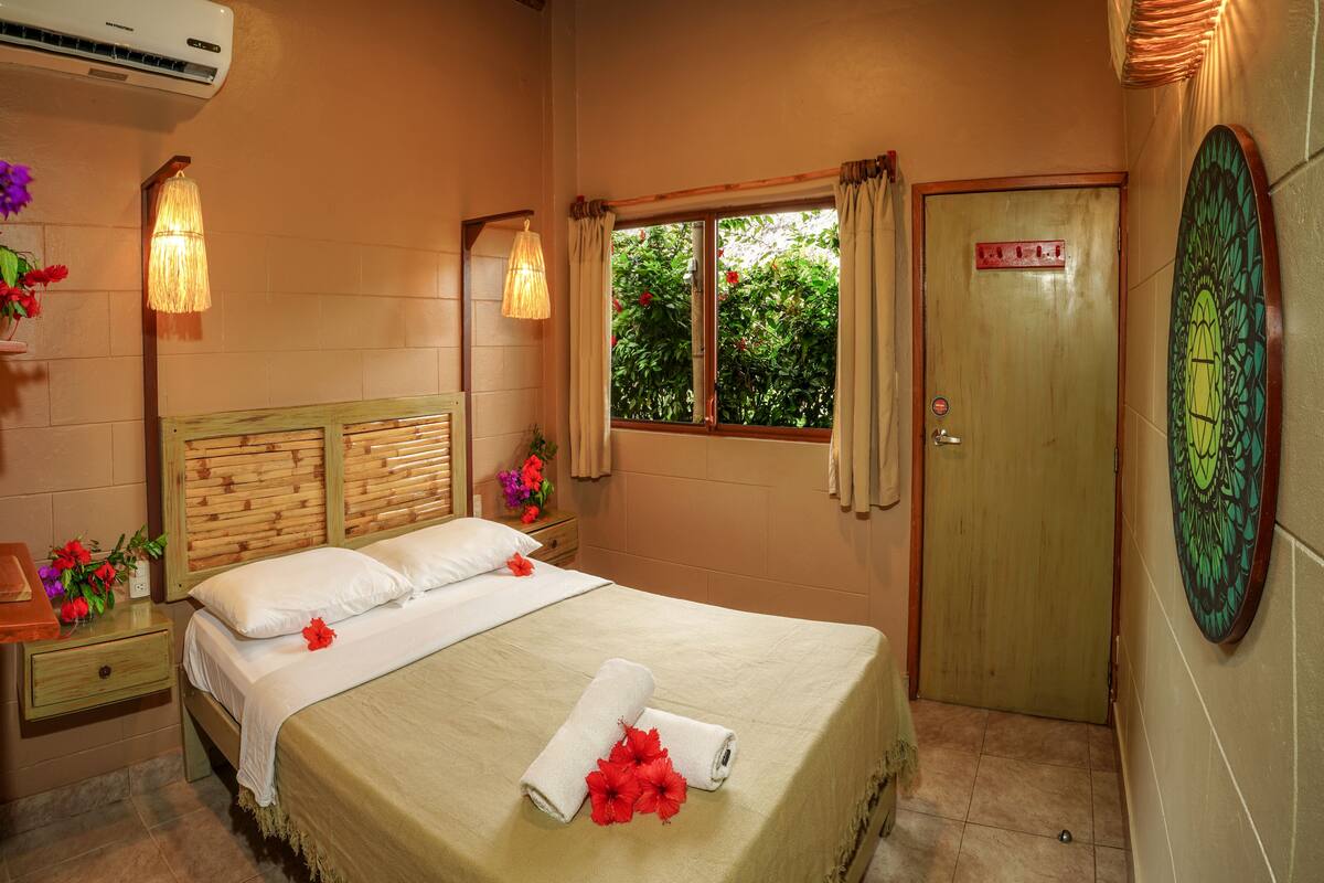 Habitación de hotel con cabecera de madera, cama con ropa blanca, ventana con cortinas y puerta con un letrero.