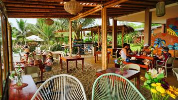 Enjoy a casual dining experience in our outdoor space, featuring wicker chairs and wooden tables, sheltered by a bamboo roof.