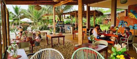 Un restaurante al aire libre con estructuras de bambú, mesas y sillas, rodeado de follaje y palmeras.