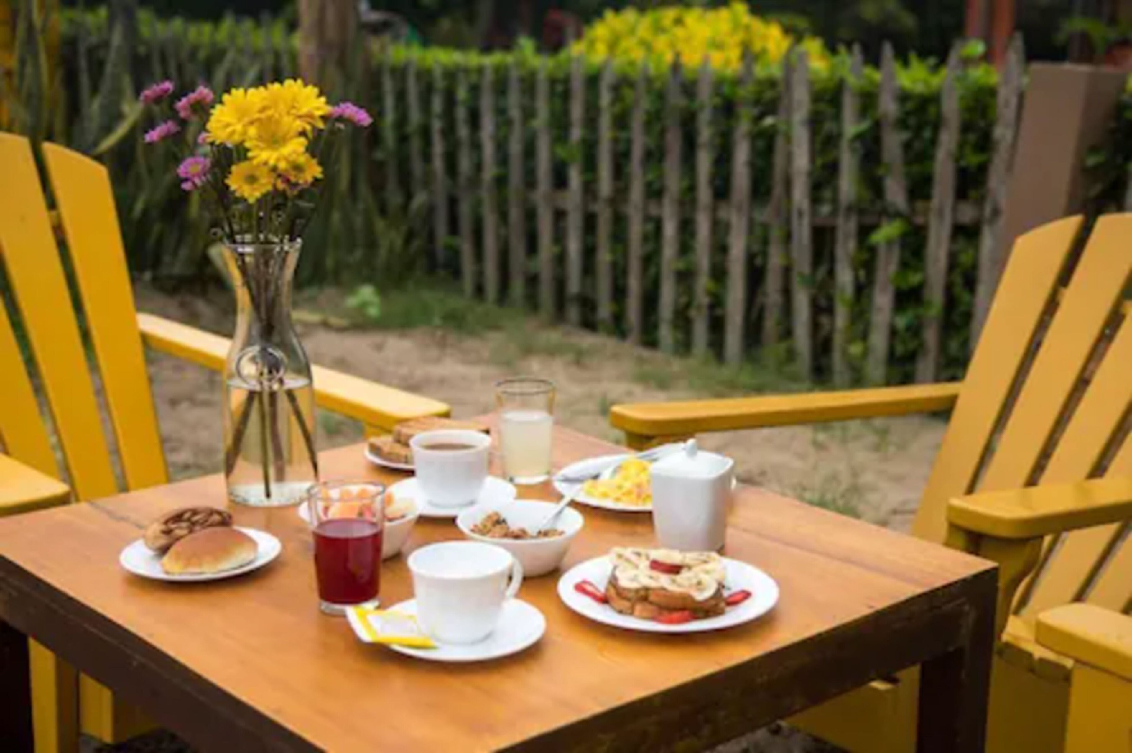 Uma mesa de madeira posta com itens de café da manhã, um vaso com flores amarelas e cadeiras amarelas em ambiente externo.