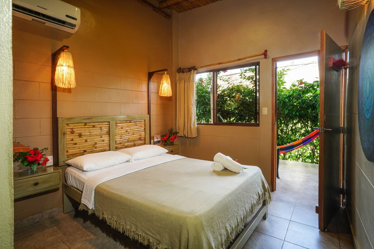 Un dormitorio con cabecera de madera, una cama con ropa de cama blanca, una mesita de noche con lámpara y una ventana con vista a la vegetación.