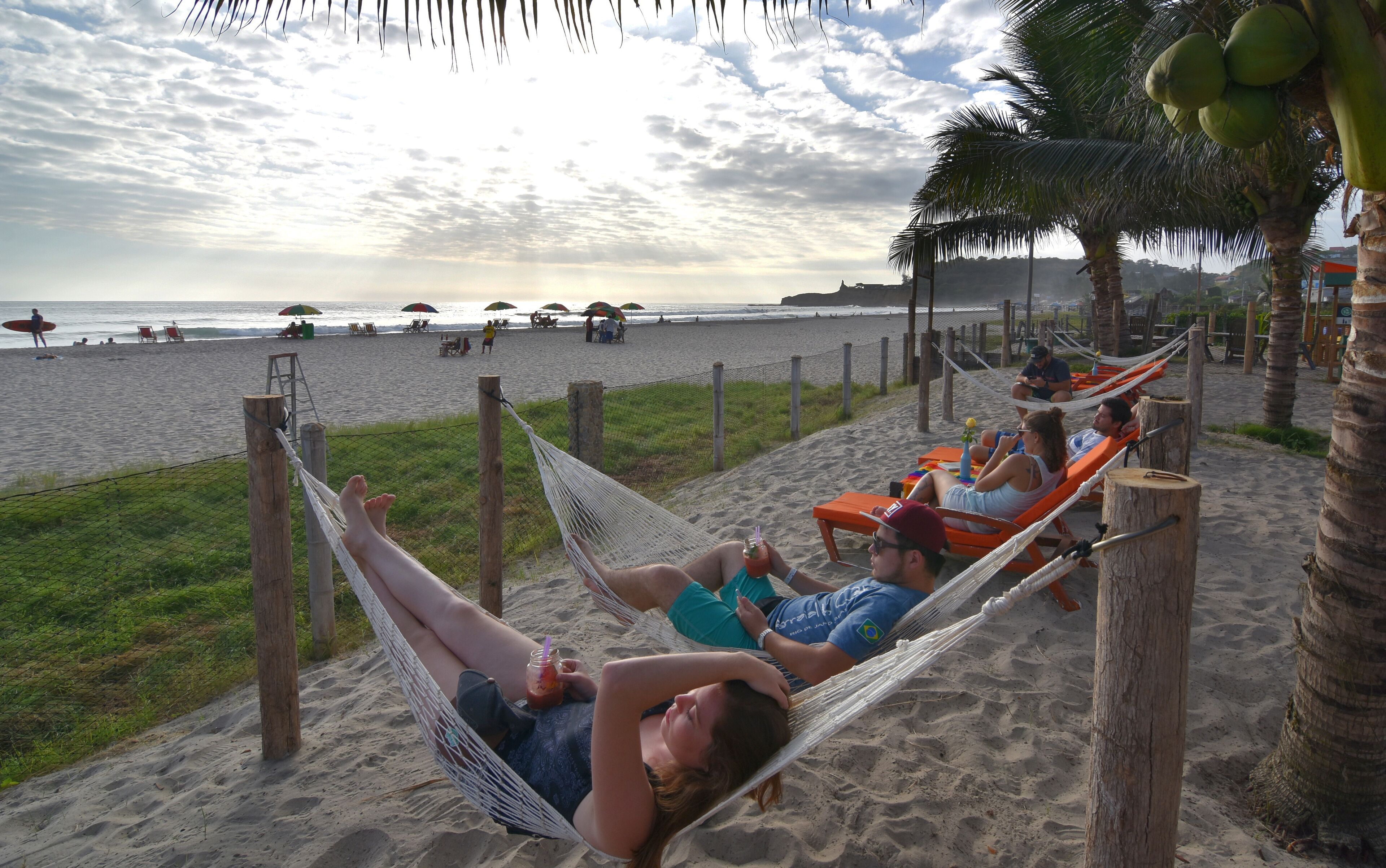 Escena de playa con gente descansando en hamacas y sillas, una valla y palmeras.