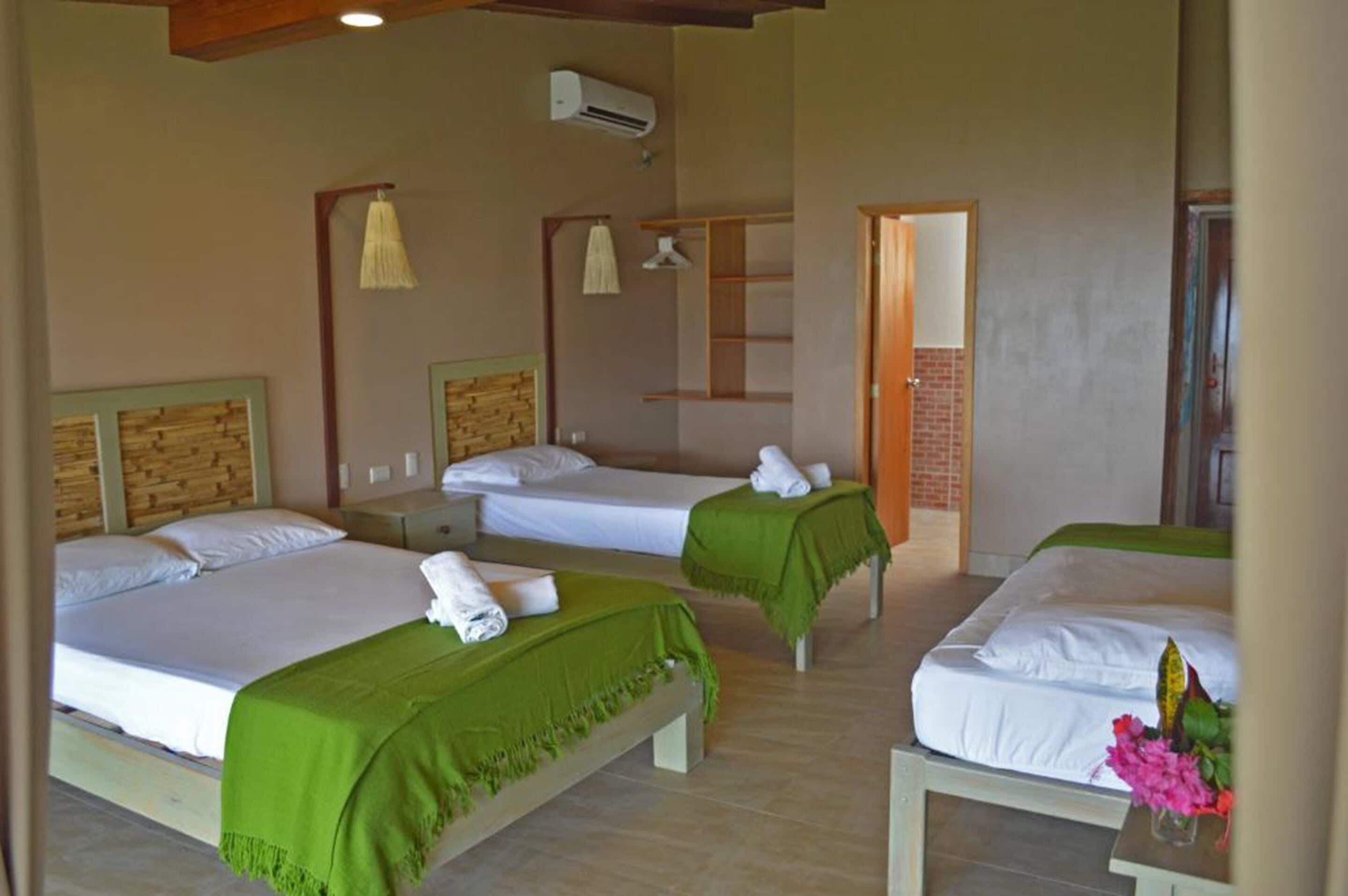 Een hotelkamer met drie eenpersoonsbedden, elk met een groene plaid en witte lakens. Elk bed heeft een houten hoofdbord, een klein nachtkastje en een wandgemonteerde airconditioning.
