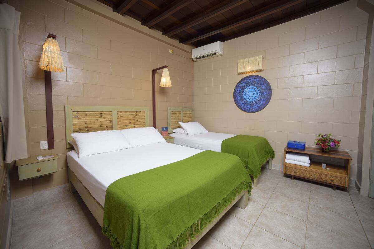 Habitación de hotel con dos camas, cabecera de madera, aire acondicionado de pared, un plato decorativo y una mesita de noche de madera con un florero.