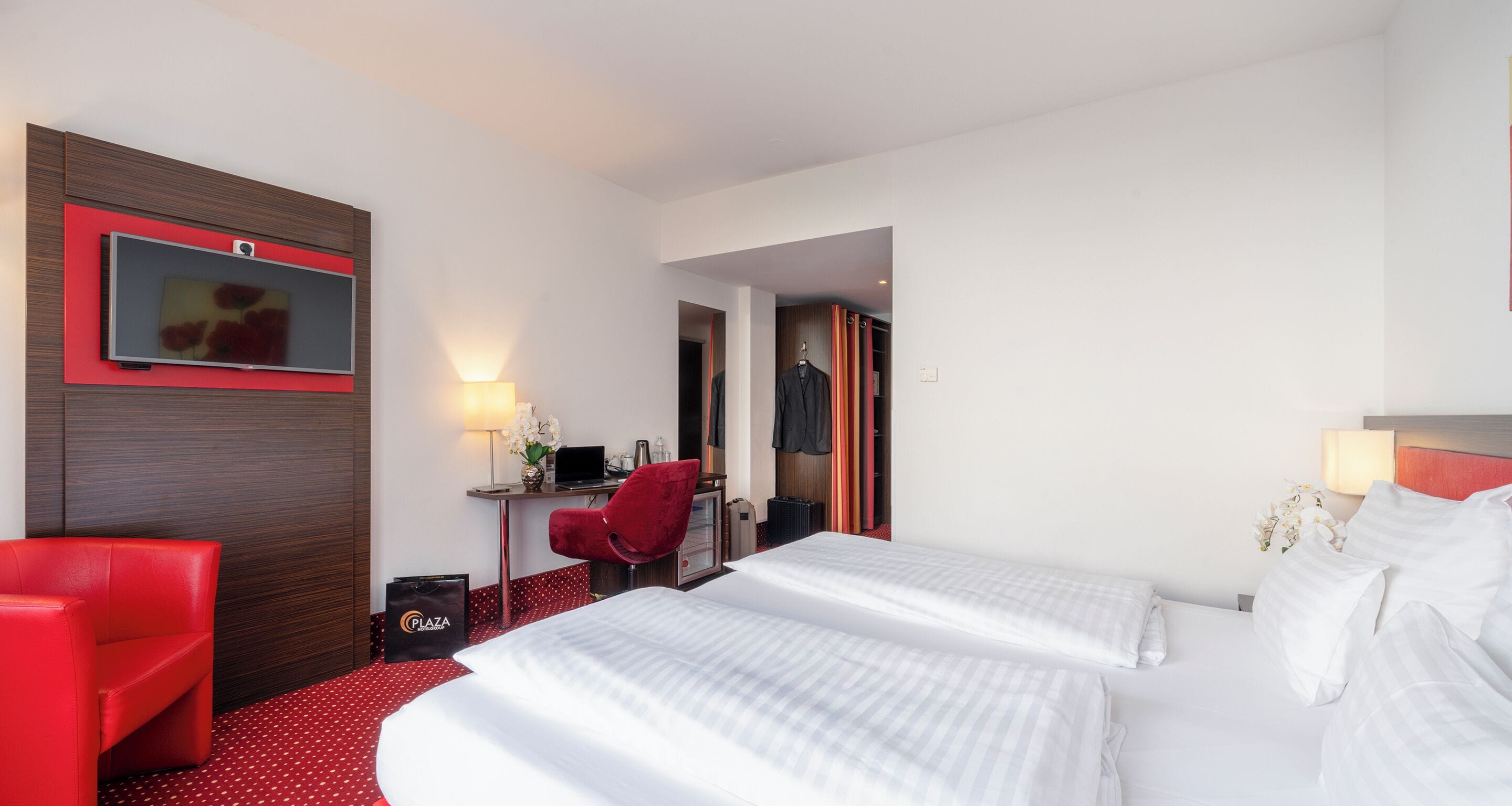 A hotel room with two beds, a desk with a computer, a red chair, and a TV mounted on the wall.