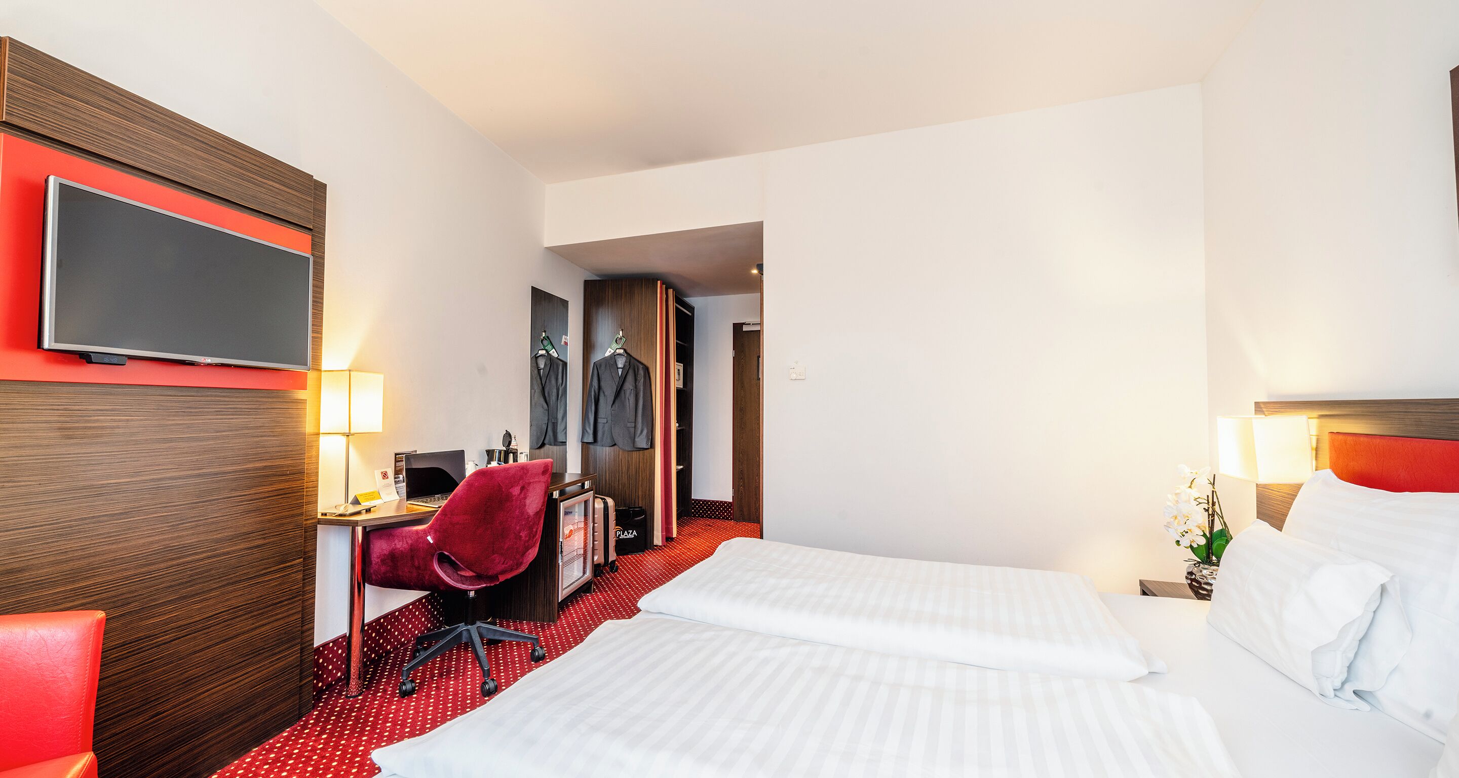 A hotel room with two beds, a desk with a computer, a red chair, and a TV mounted on the wall.