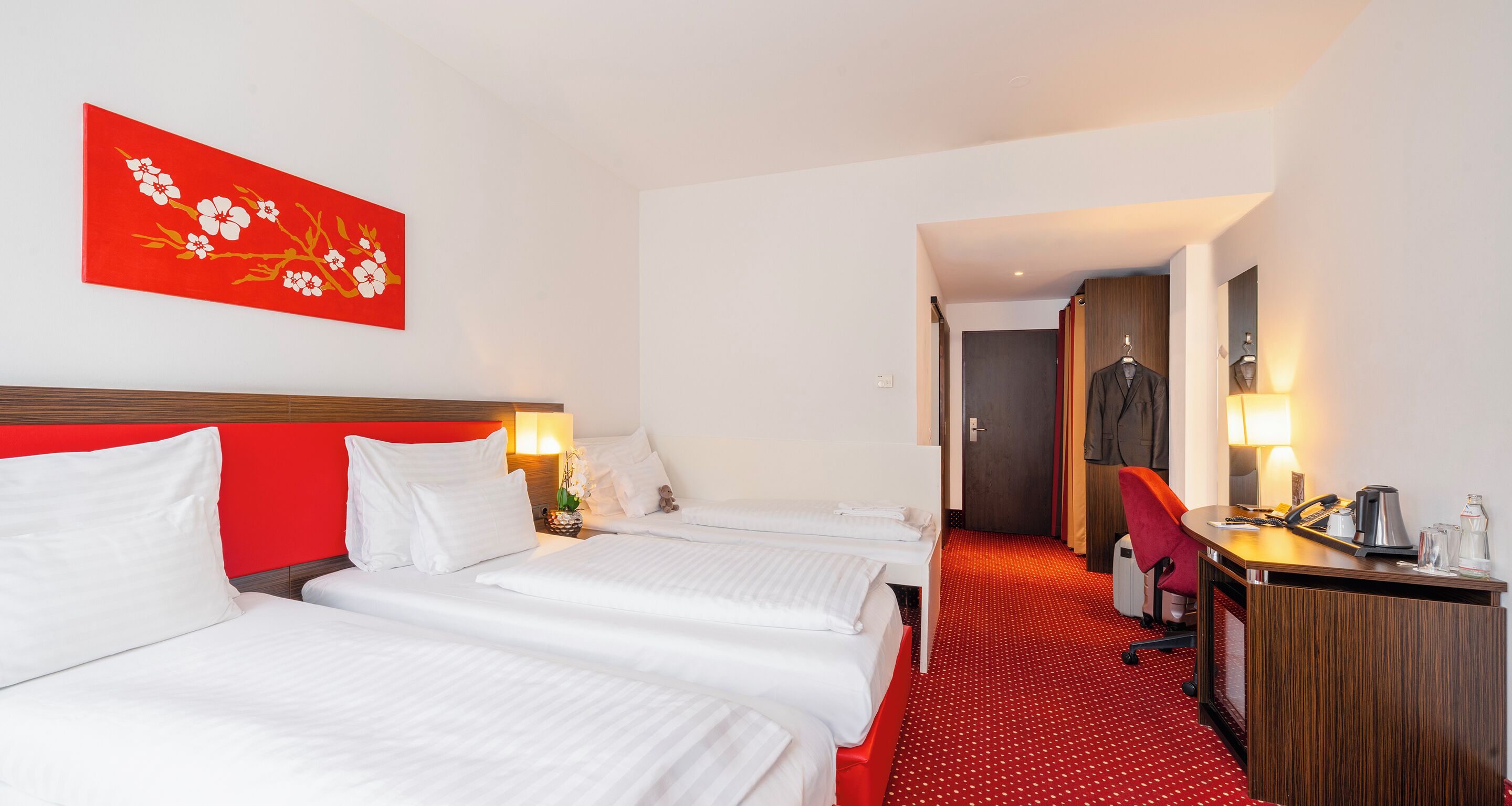 A hotel room with two beds, a red wall with a floral painting, a desk with a chair, and a door leading to a bathroom.