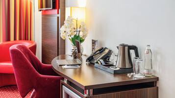 A hotel room with a wooden desk, a red chair, a laptop, a telephone, a coffee maker, a vase with white flowers, and a bottle of water.